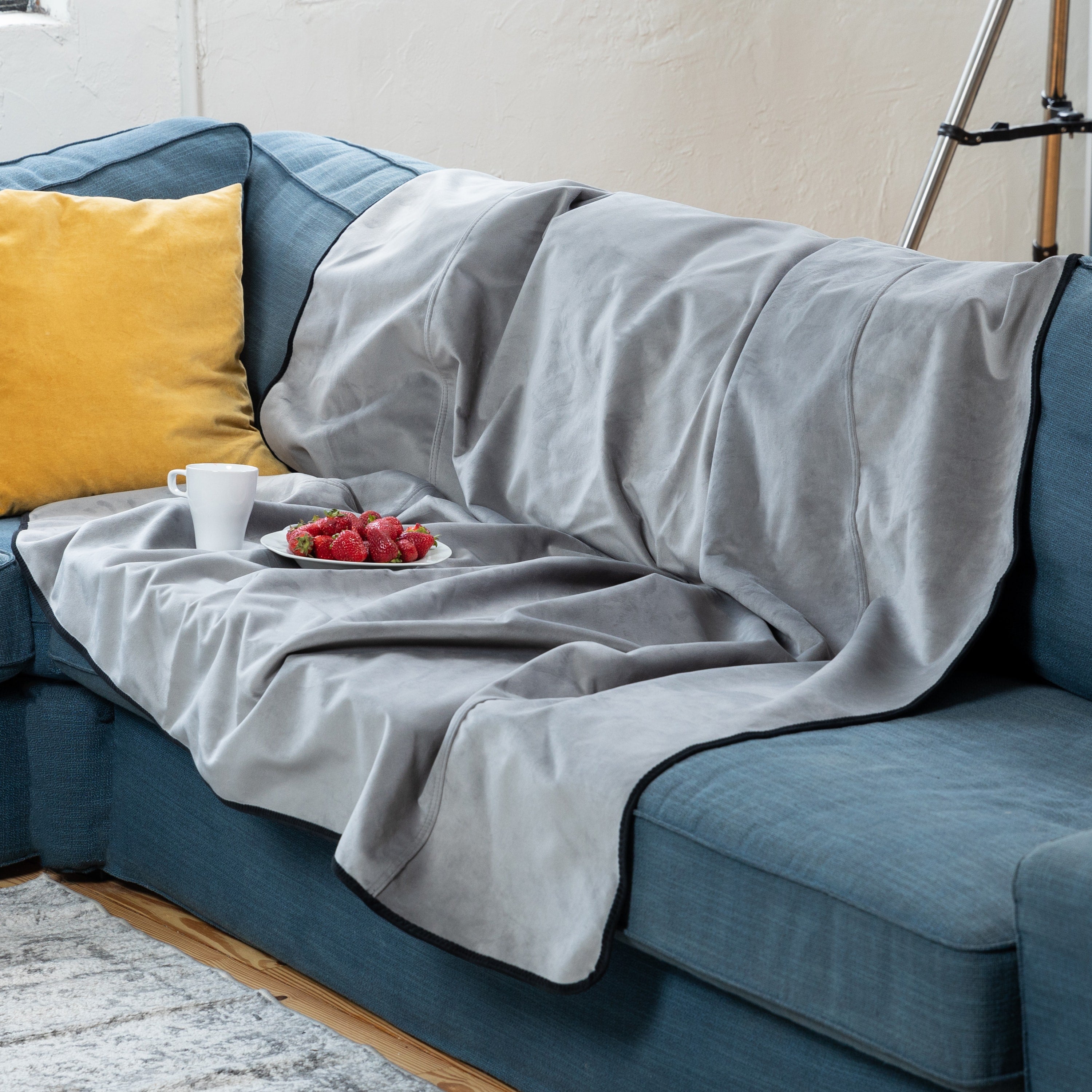 Gray blanket draped over a blue sofa with a yellow pillow and a plate of strawberries on a white tablecloth.