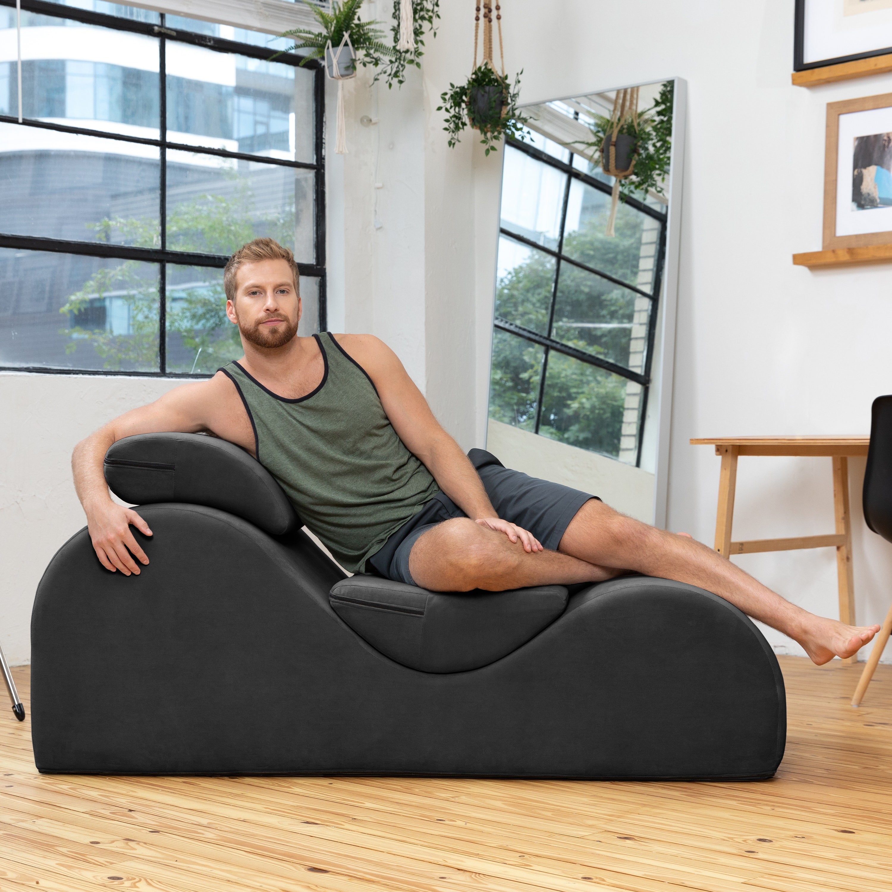 A man wearing a green tank top and dark shorts reclines on a modern, curved black foam lounger in a bright, contemporary loft-style room with wood floors and large windows.