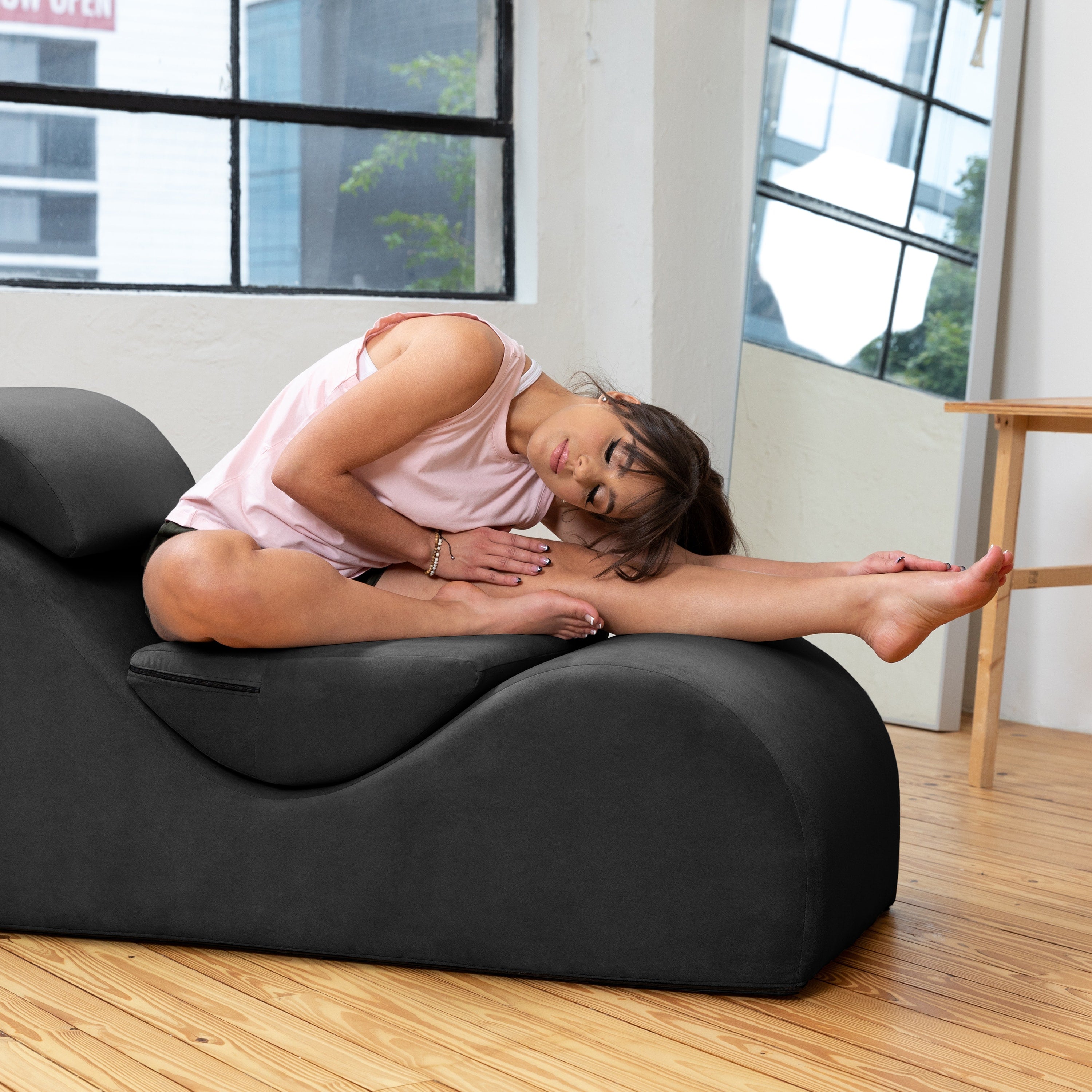 A woman in a pink tank top sits on a black molded lounge chair in a bright room with wood floors and large windows.
