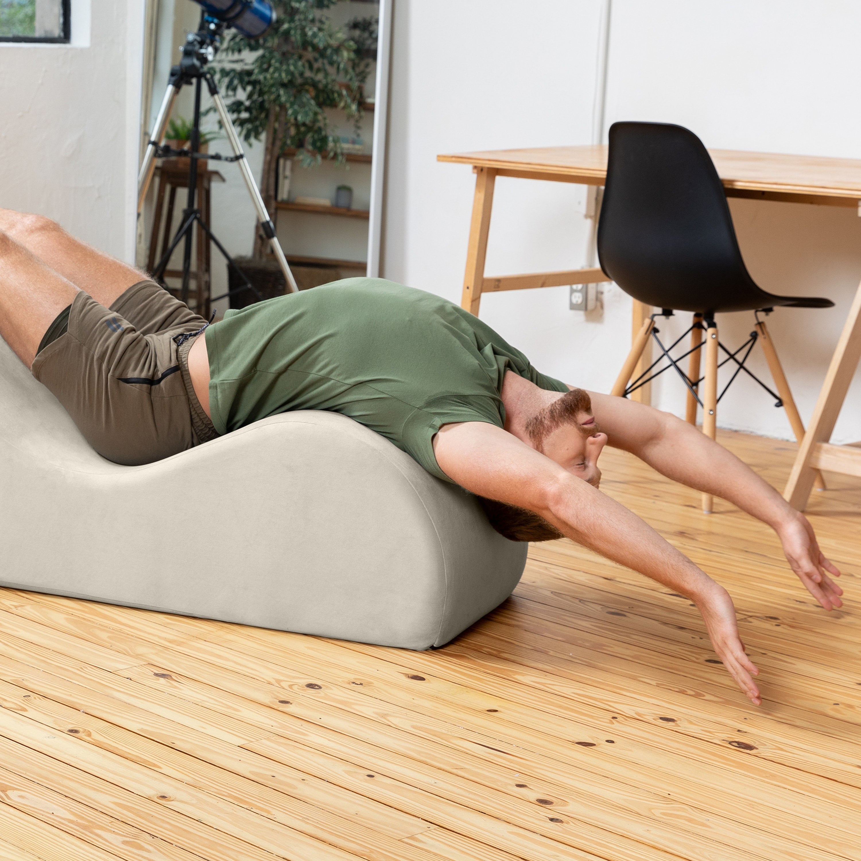 A man in an olive green t-shirt and brown shorts lies stretched out on a curved, light beige foam massage or chiropractic table on a light wood floor in a room with a wooden dining table and black chair.