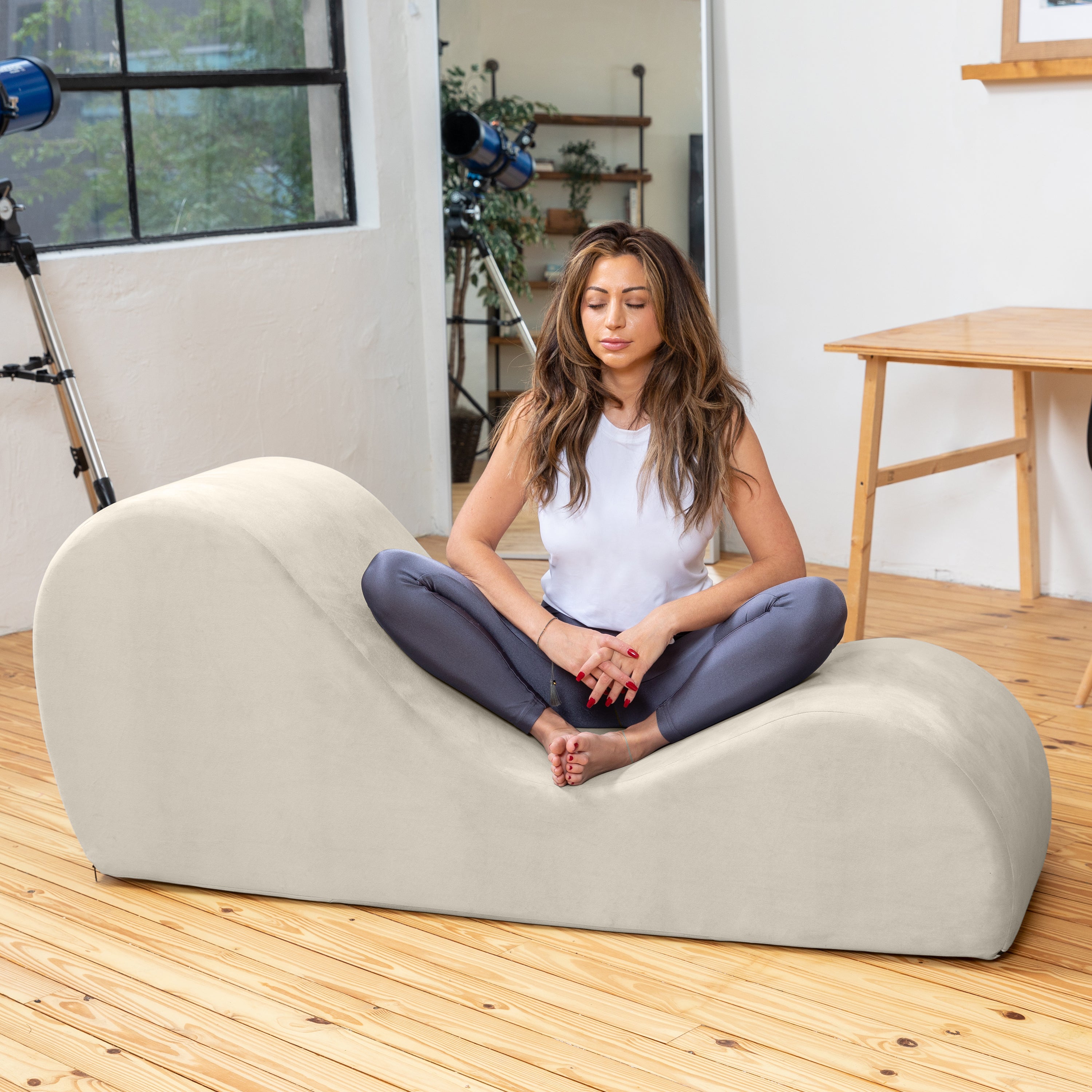 Woman sitting on a beige bean bag chair in a room with a window and wooden floor.