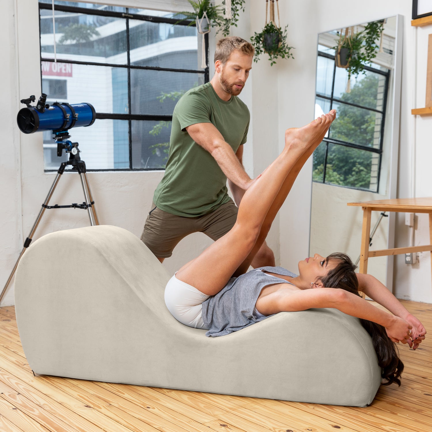 Person receiving leg stretch from another person on a beige curved foam roller in a room with large windows and plants.