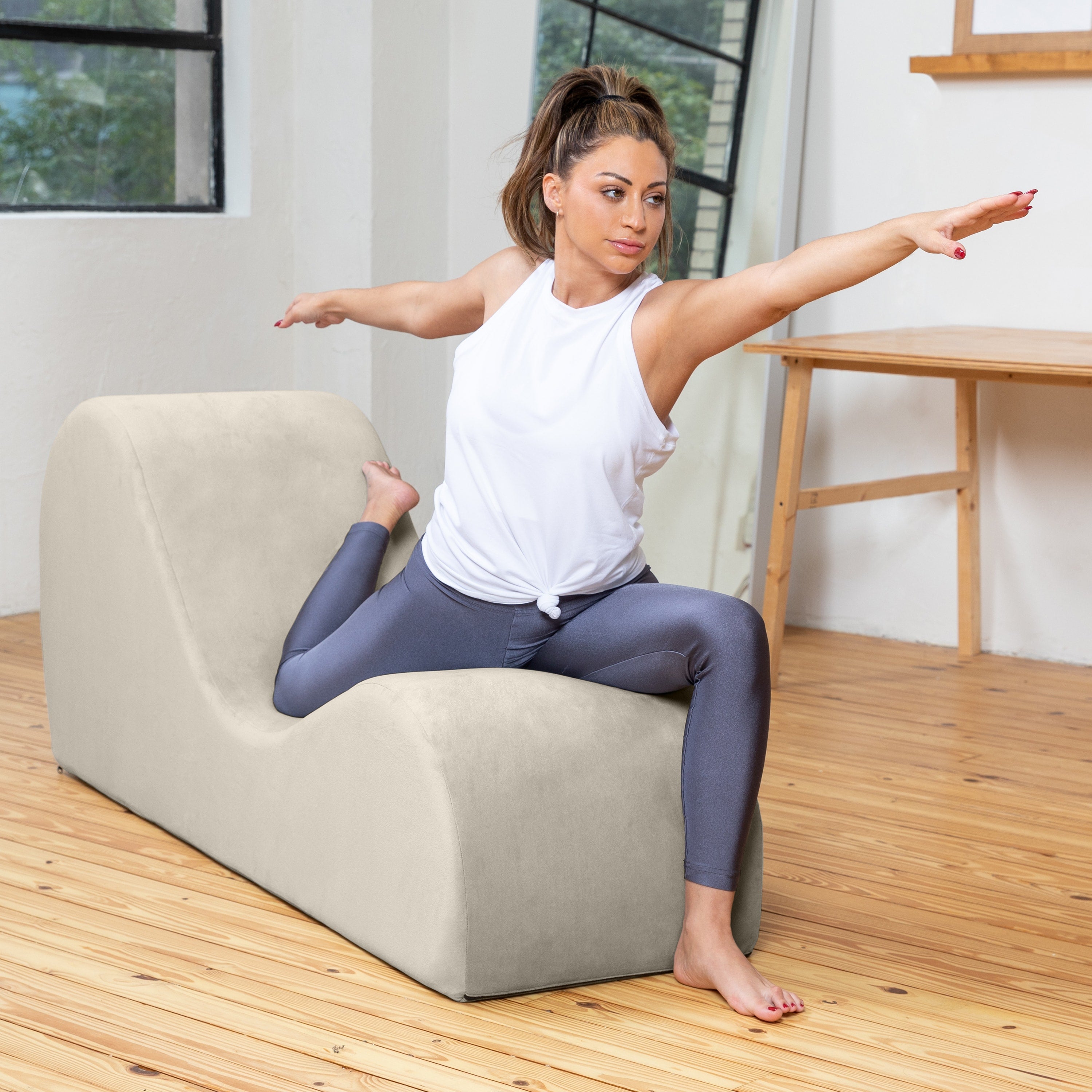 A woman in a white sleeveless top and slate blue leggings sits on a beige upholstered lounge chair in a bright room with wood floors, holding her leg in a yoga pose.