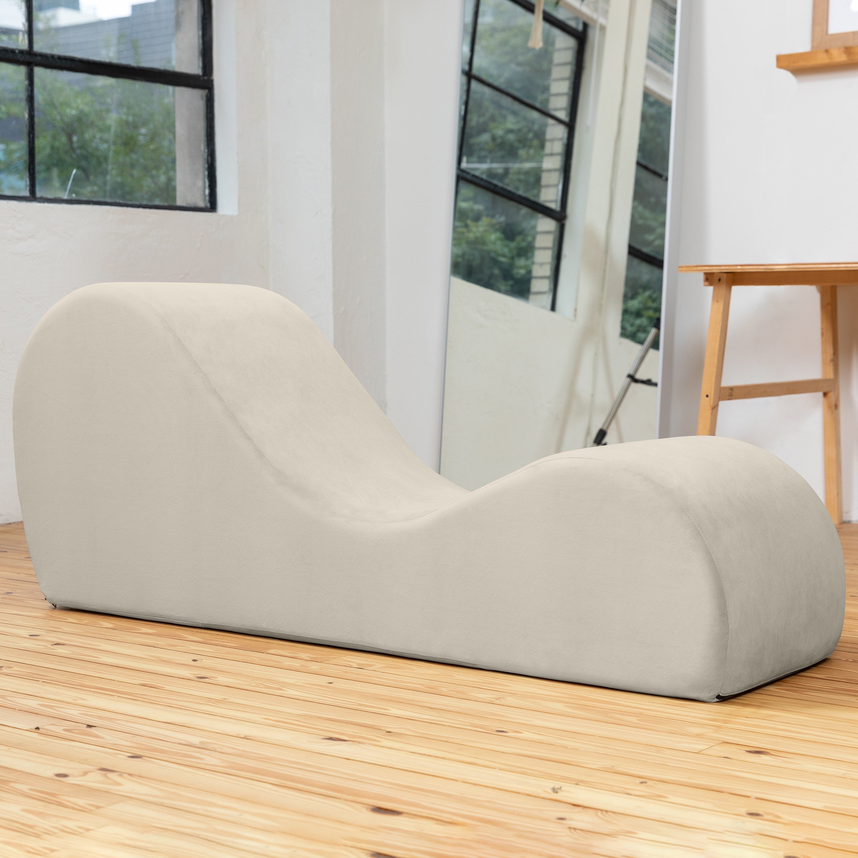 A modern curved lounge chair in a light beige fabric, placed on a light wood floor in a bright room with a mirror, window, and wooden table visible in the background.
