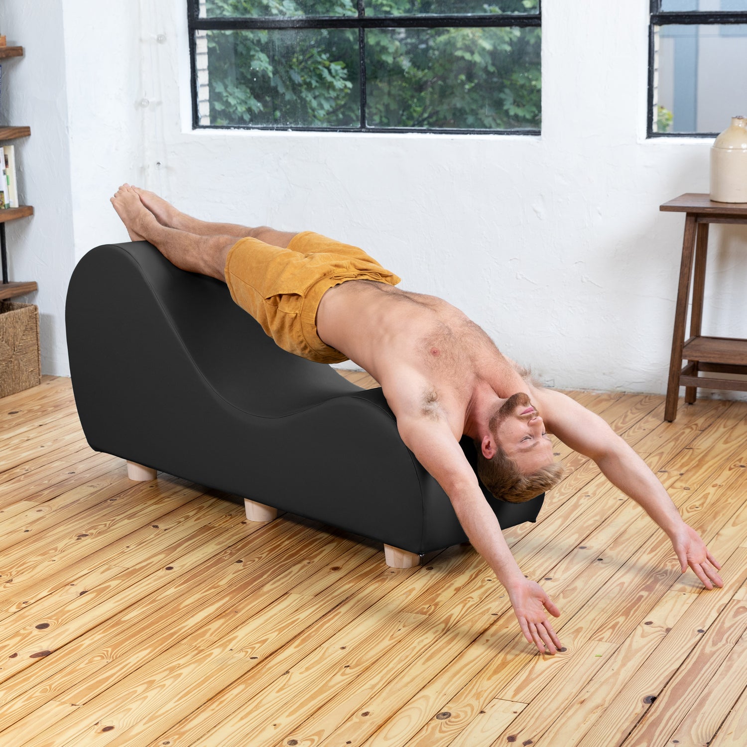 Person using a black curved exercise tool on a wooden floor with a white wall and window in the background.