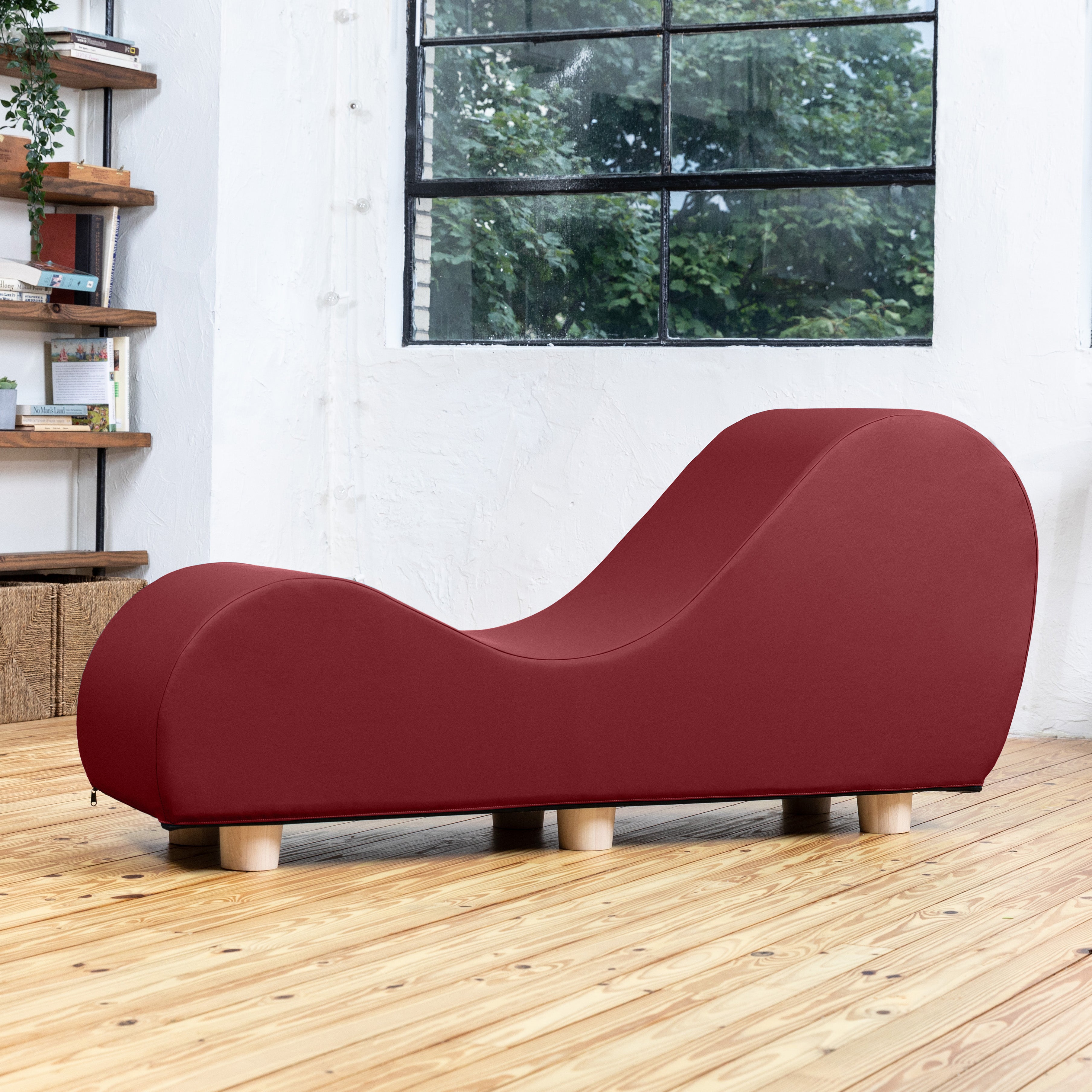 A modern curved lounge chair in deep burgundy fabric with light wood legs, positioned on a light wood floor in a bright room with a window and bookshelf.