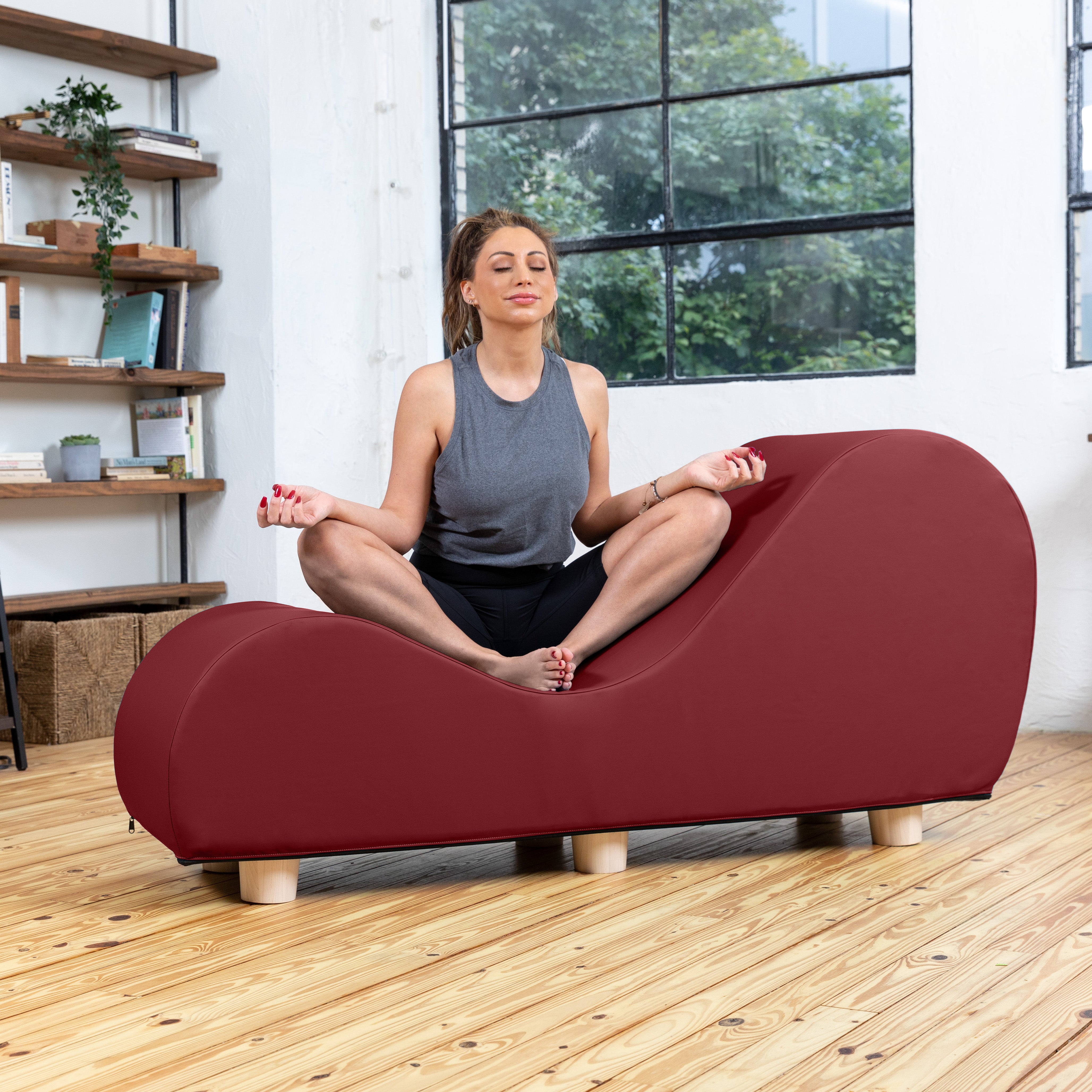 A woman in a gray tank top and black pants sits cross-legged in a meditative pose on a curved, cushioned floor pad in a bright room with wood floors and a large window.
