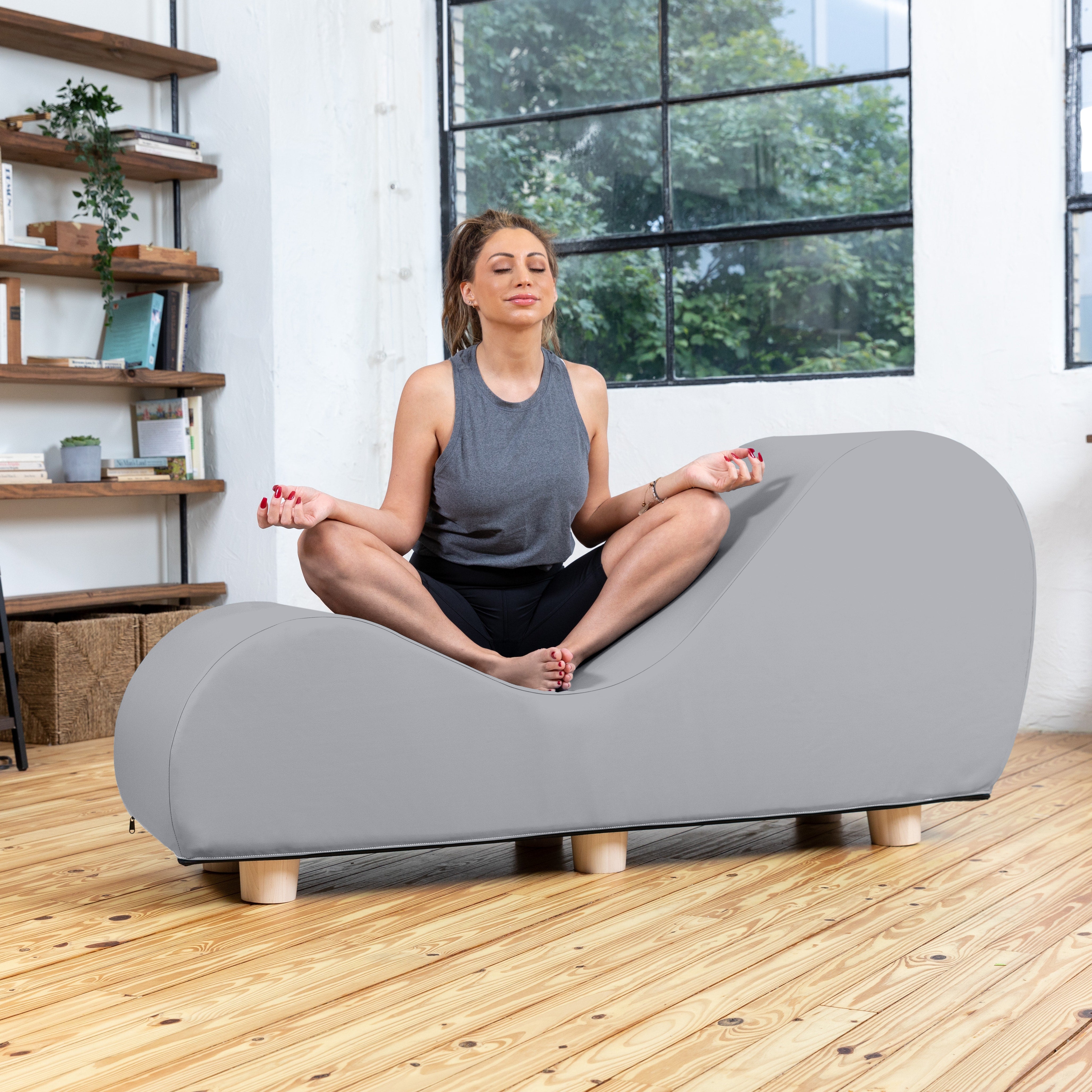 Woman sitting in a meditative pose on a modern gray sofa in a bright room with large windows.