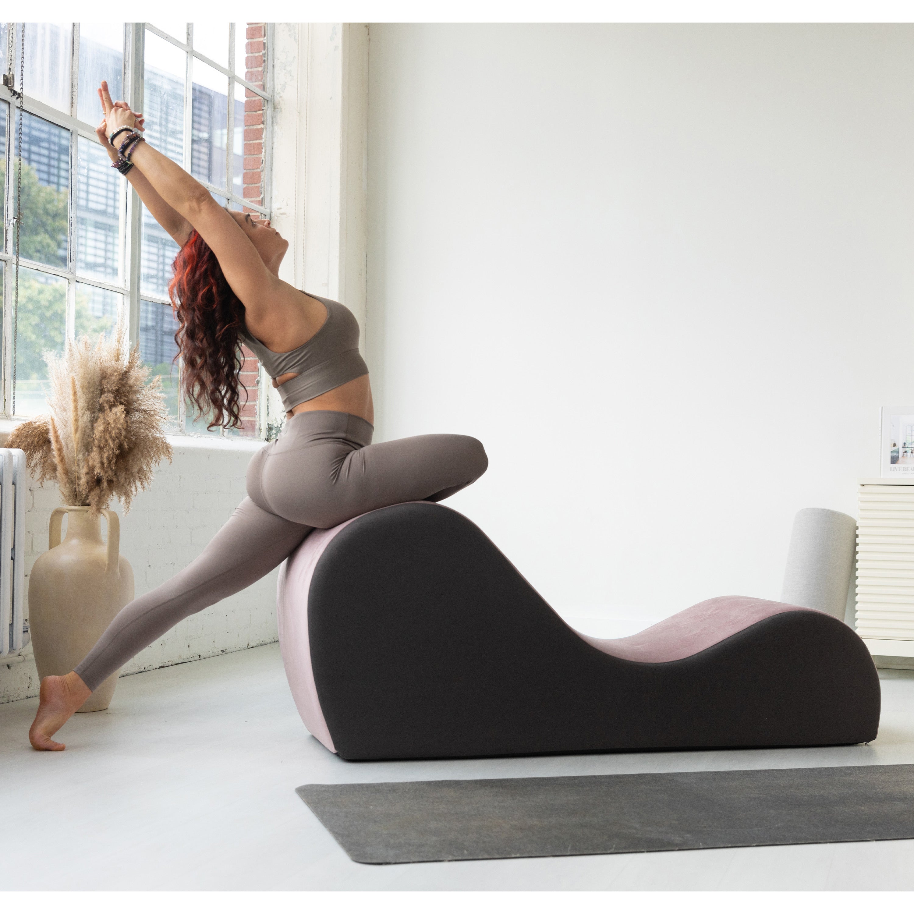 Woman using a yoga chaise in a bright room with large windows.