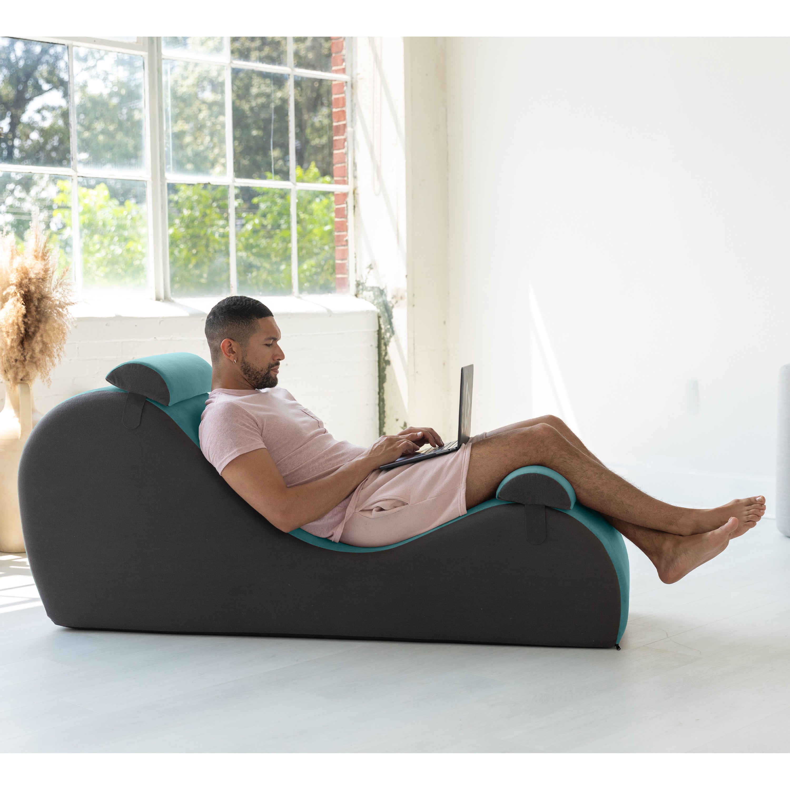 Man using a laptop on a modern gray foam chair in a bright room with large windows.