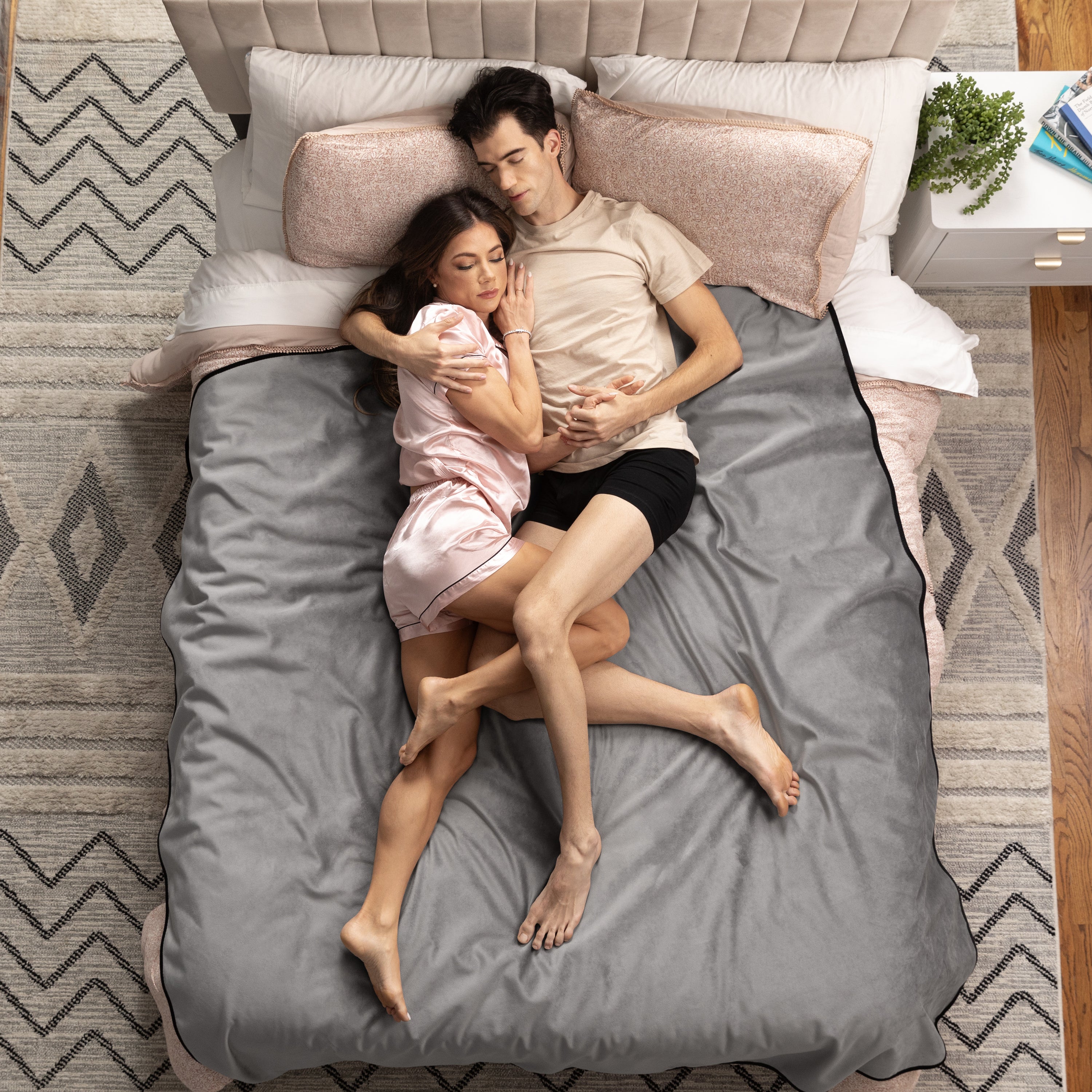 Couple lying in bed under a gray throw on a patterned rug.