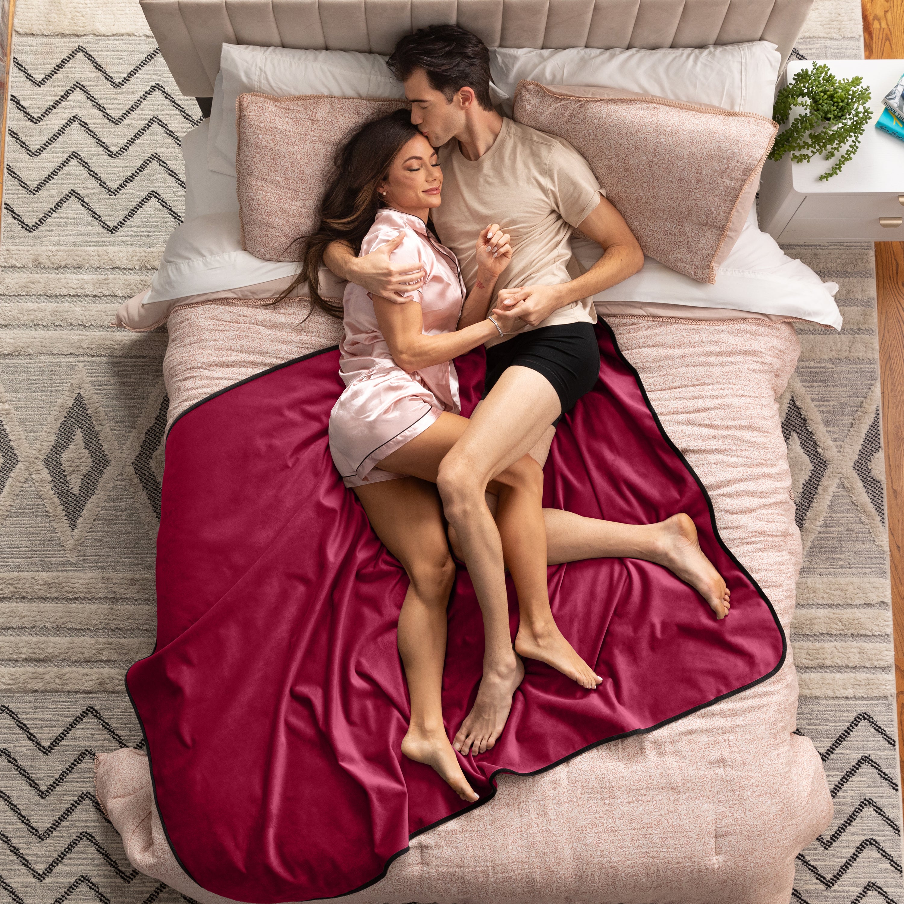 Couple lying on a bed under a red blanket in a bedroom setting.