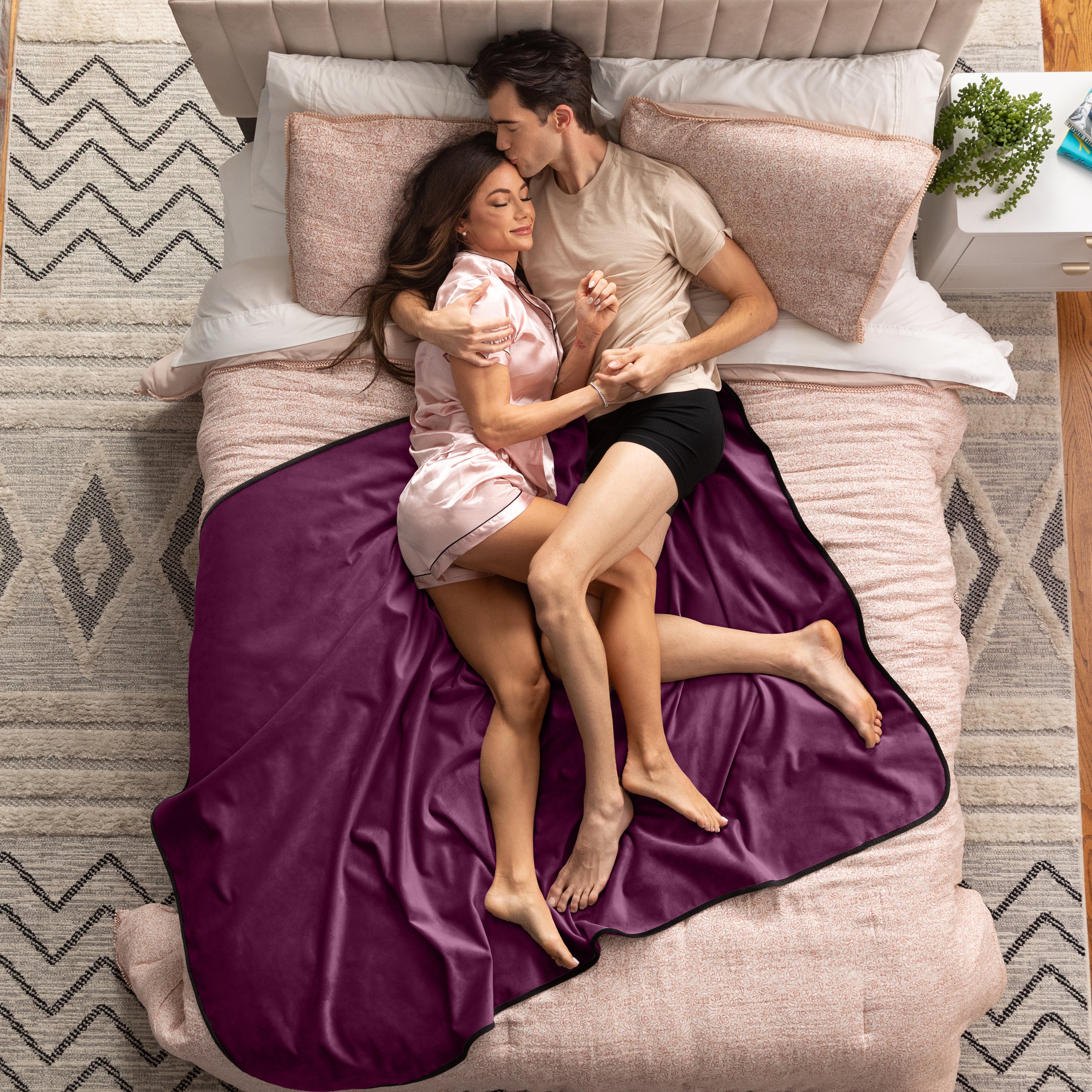 Couple lying on a bed under a purple blanket in a bedroom setting.