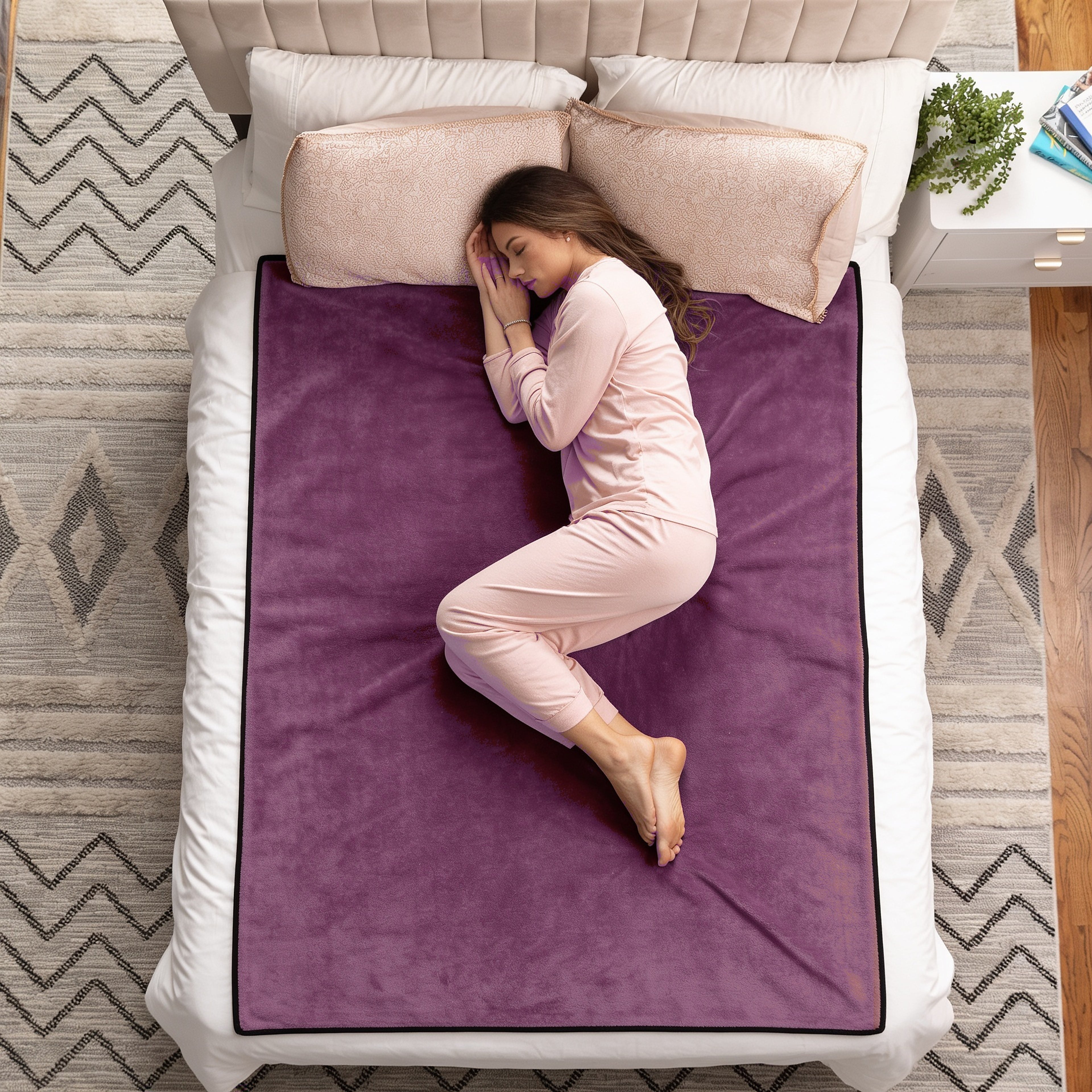 A woman in light pink pajamas sleeps curled up on a purple fleece blanket spread across a white bed, shown from a top-down view in a bedroom setting.