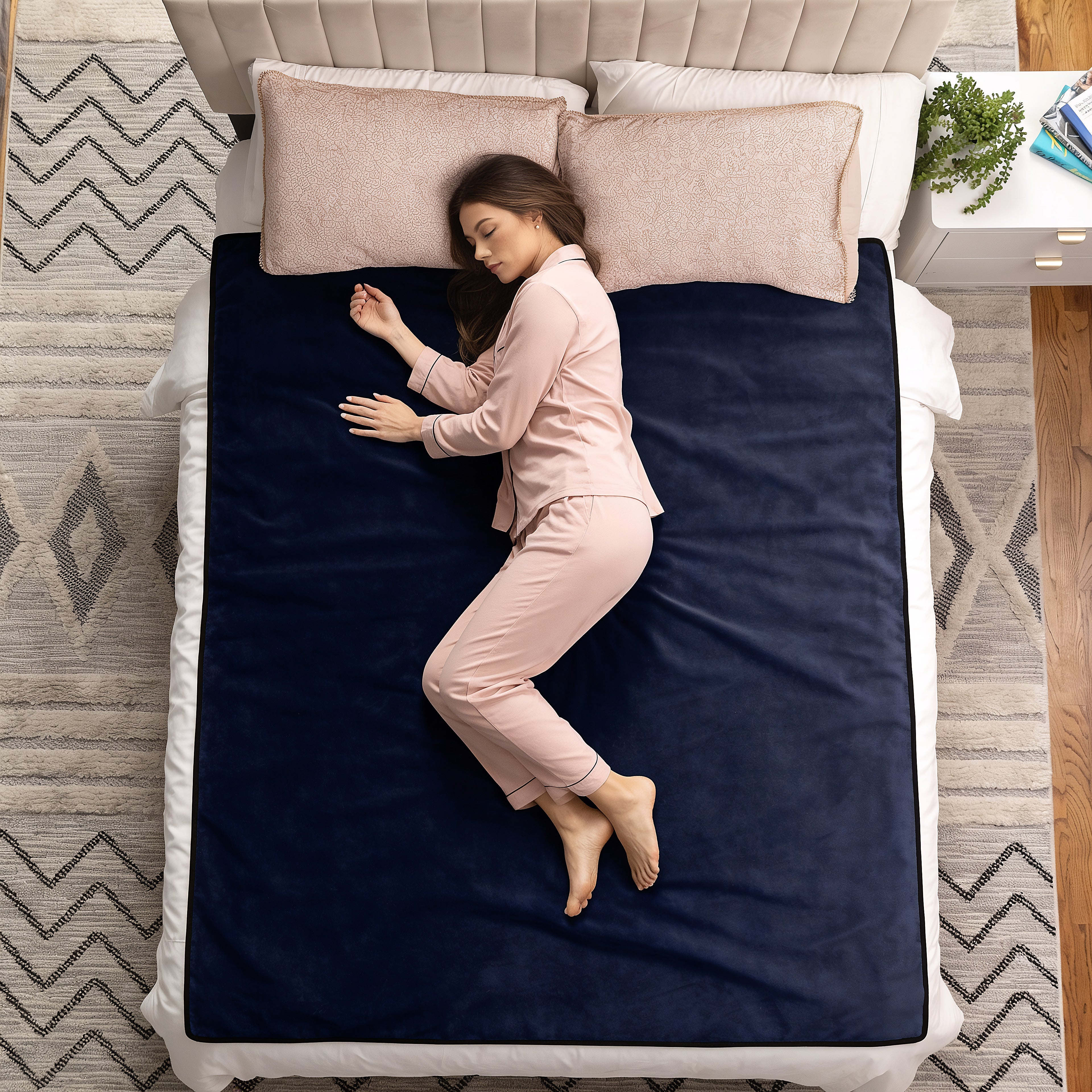 A woman in light pink pajamas lies on her side on a bed with a dark navy blue blanket or mattress cover, white bedding, two blush pink pillows, and a patterned rug in a bedroom setting.