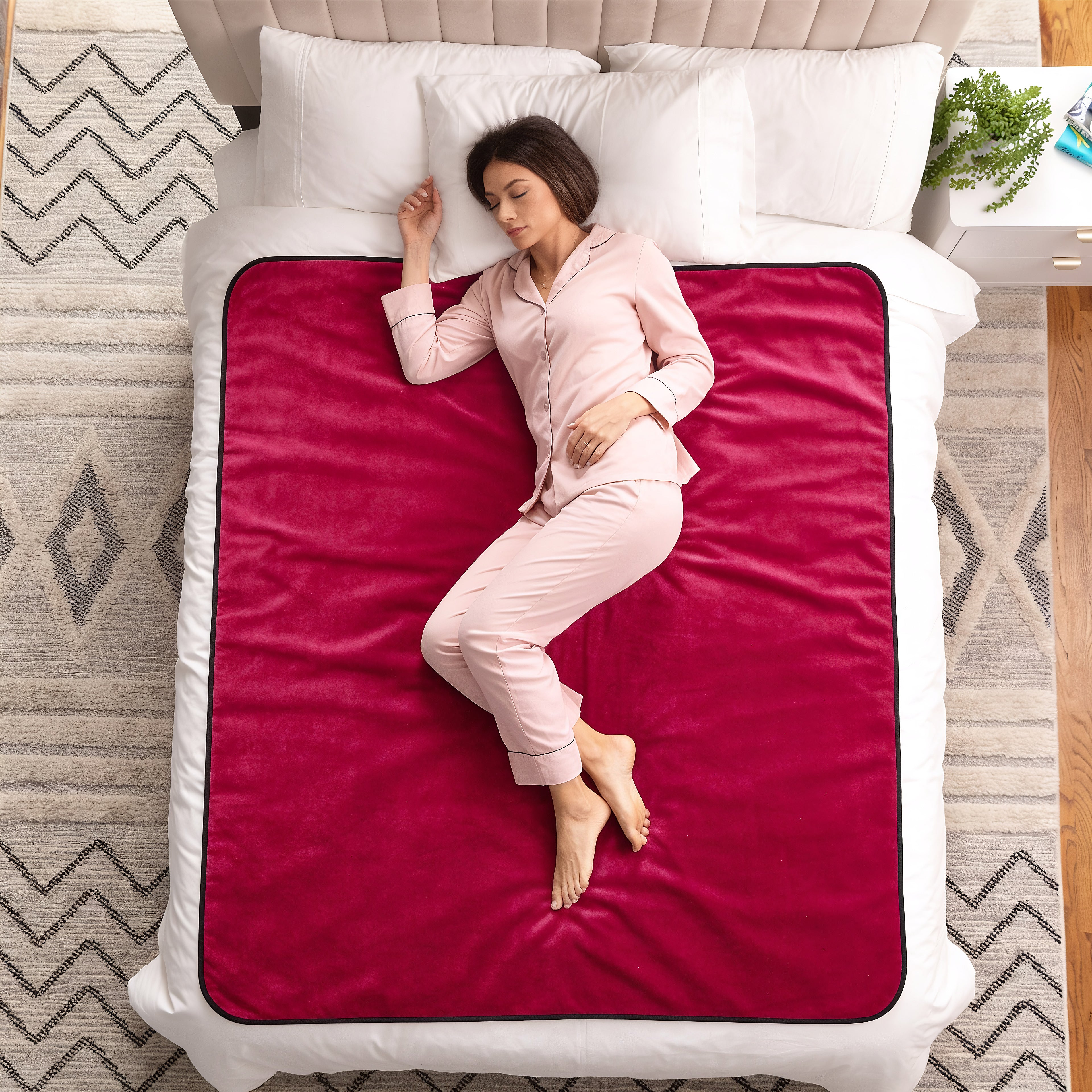 A woman in light pink pajamas lies on her side on a bed, showing a plush magenta-pink blanket or comforter with a black border over white bedding. The scene is photographed from above on a patterned rug.
