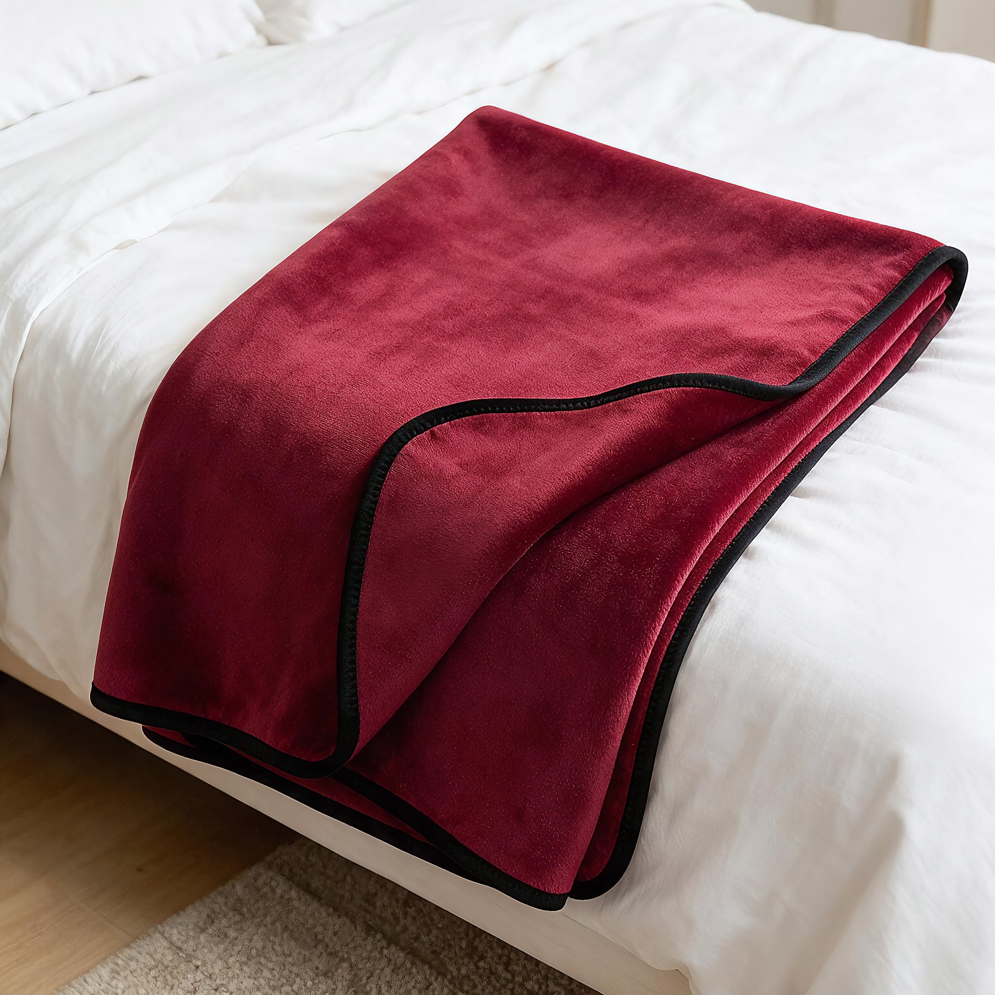 A folded burgundy fleece blanket with black trim draped over a white bed.