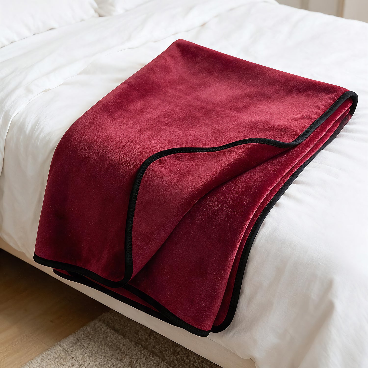 A folded burgundy fleece blanket with black trim draped over a white bed.