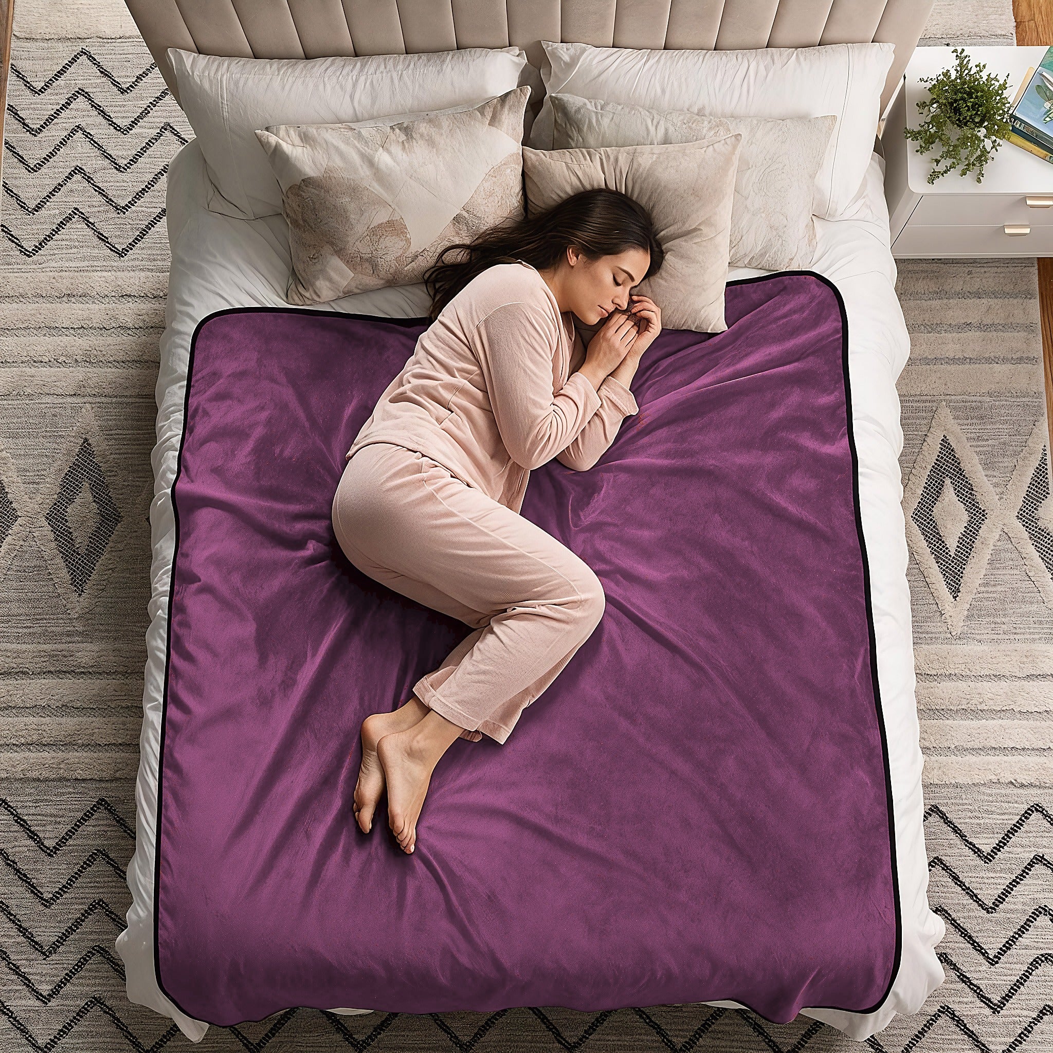 A woman in light pink pajamas sleeps curled up on a bed with a purple plush blanket spread across the mattress. The bed has white bedding, a beige upholstered headboard, and a patterned gray rug in a bedroom setting.
