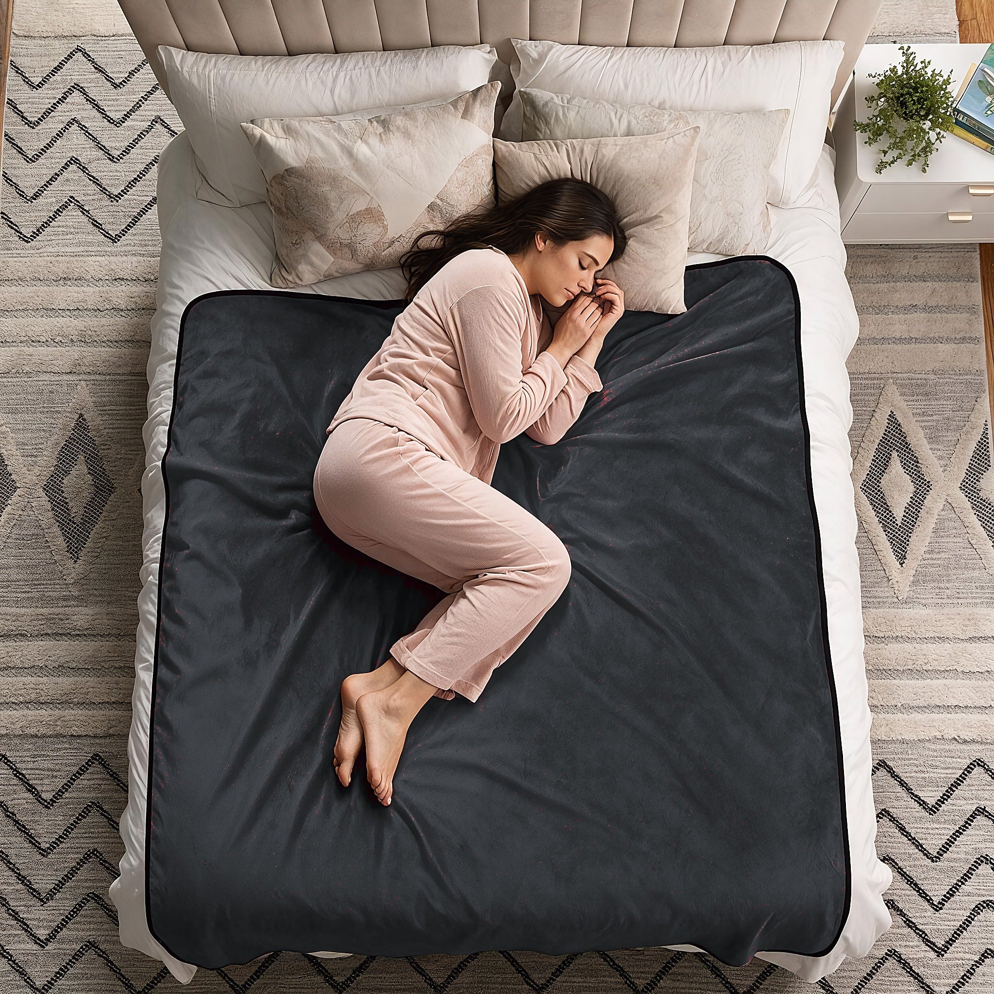 A woman in a light pink pajama set sleeps curled up on a dark charcoal gray blanket spread across a bed with white bedding and beige pillows in a styled bedroom.