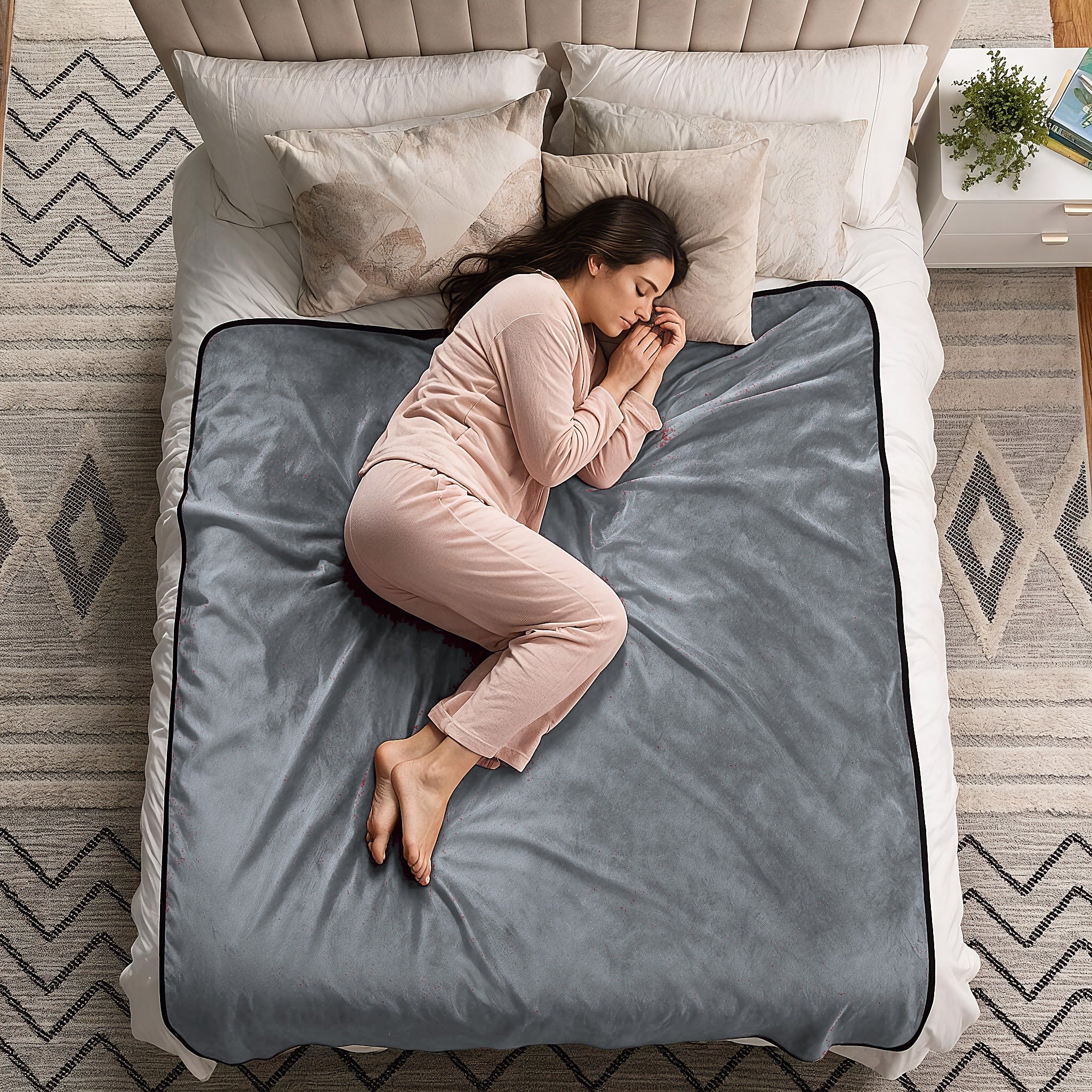 A woman in a light pink pajama set sleeps curled up on a gray blanket spread across a bed in a styled bedroom setting.