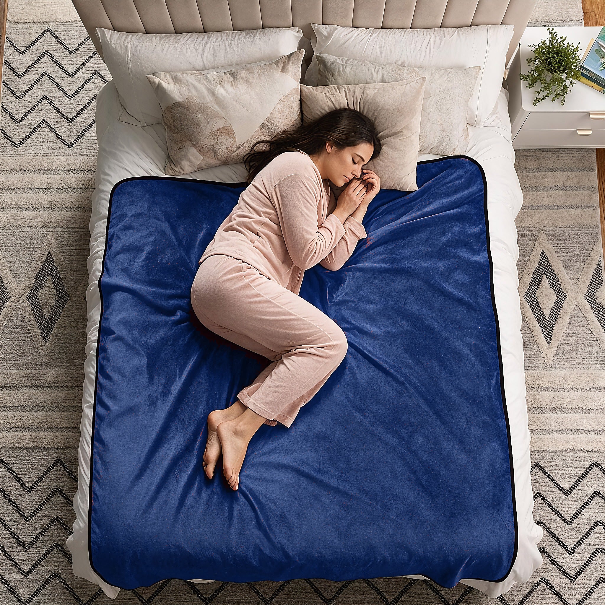 A woman in light pink pajamas sleeps curled up on a blue plush blanket spread across a white bed in a styled bedroom.