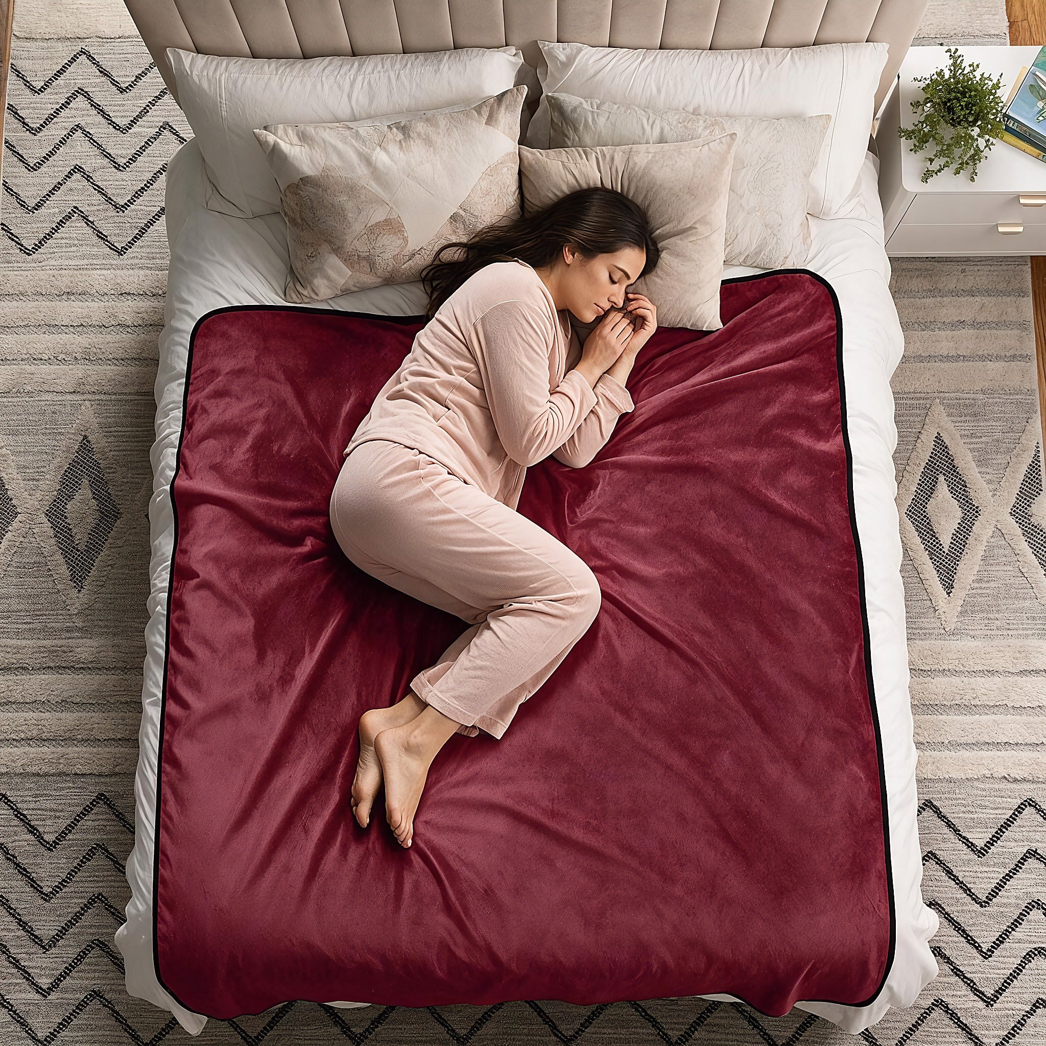 A woman in light pink pajamas sleeps curled up on a bed with a deep burgundy fleece blanket spread across the mattress, surrounded by white bedding and pillows.