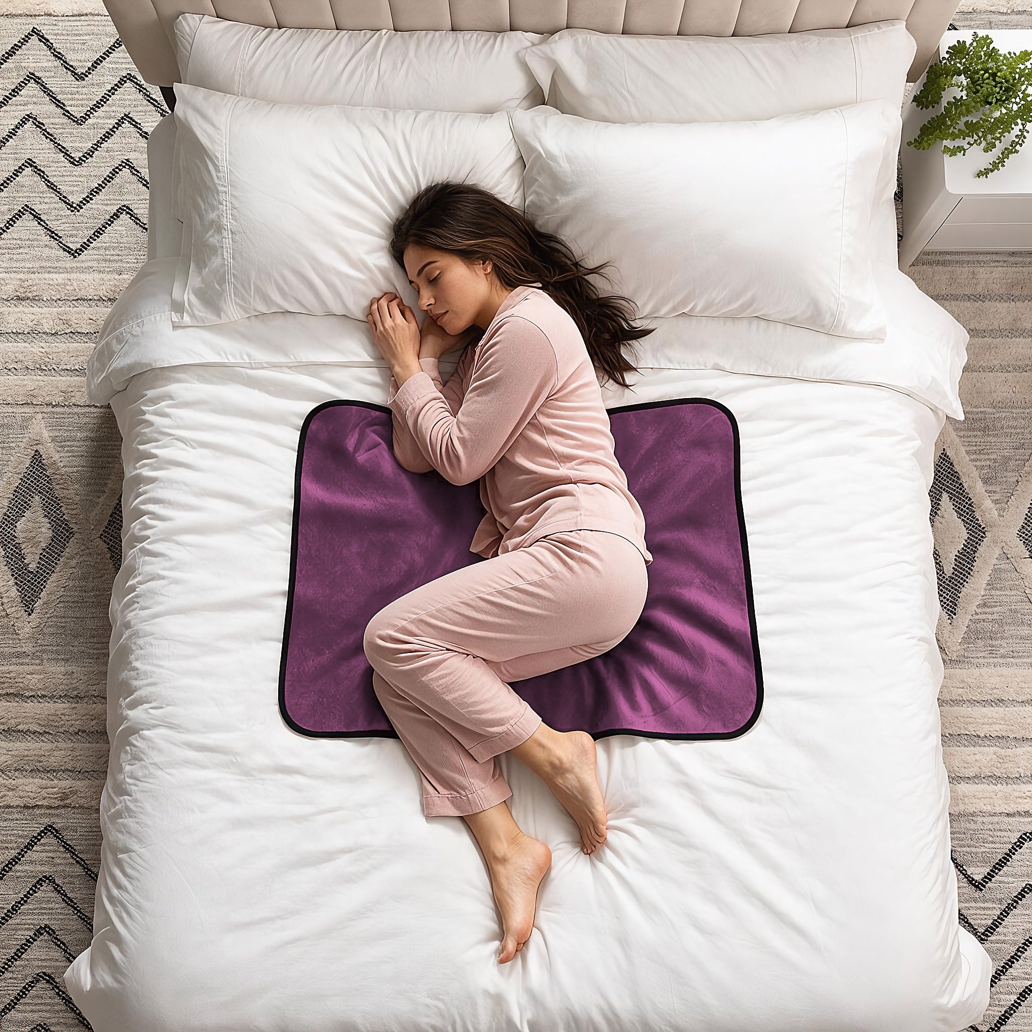 A woman in a pink pajama set sleeps curled up on a white bed, resting on a purple contoured mattress topper with a black border.