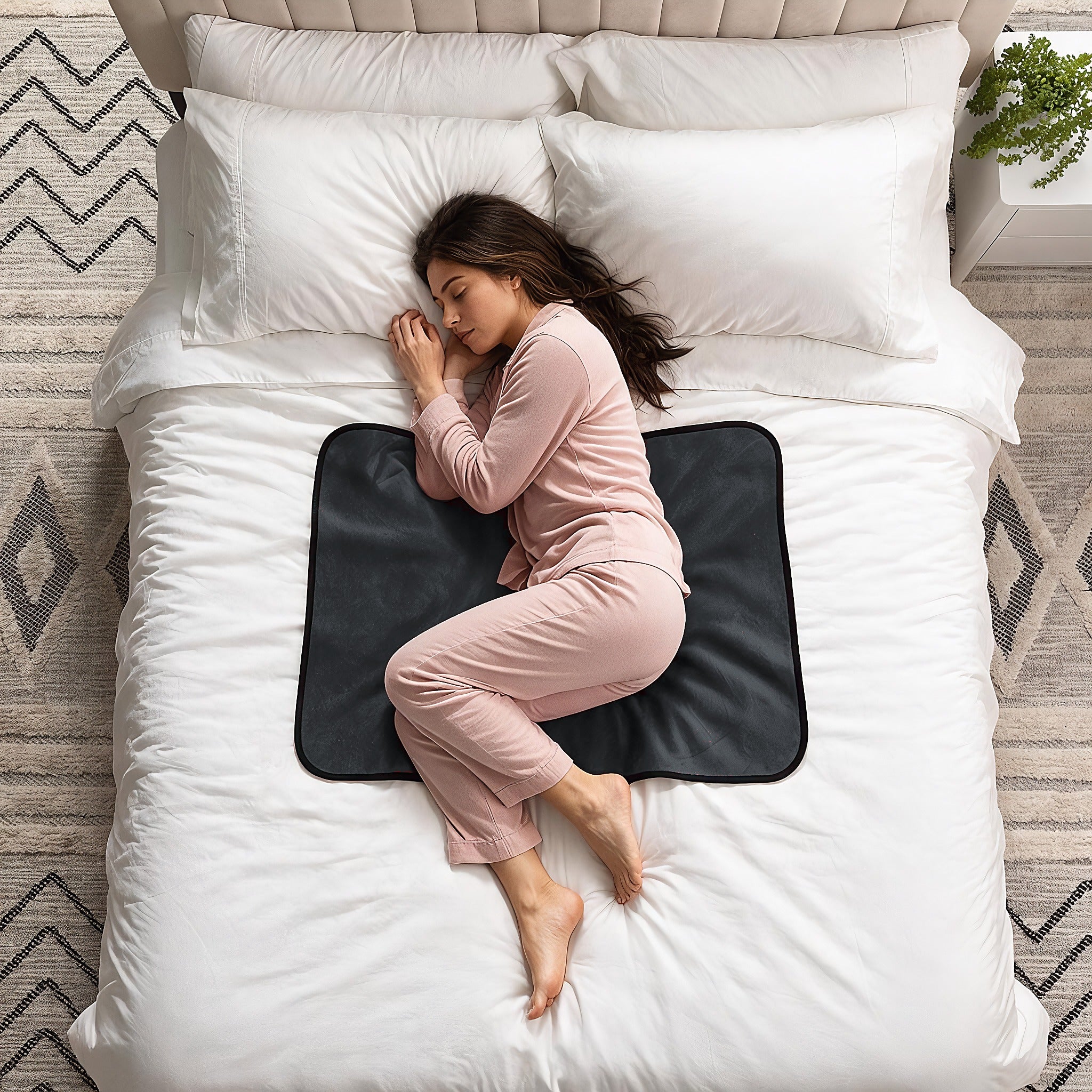 A woman in pink pajamas sleeps curled up on a black heated mattress pad on a white bed with white pillows and a patterned rug.