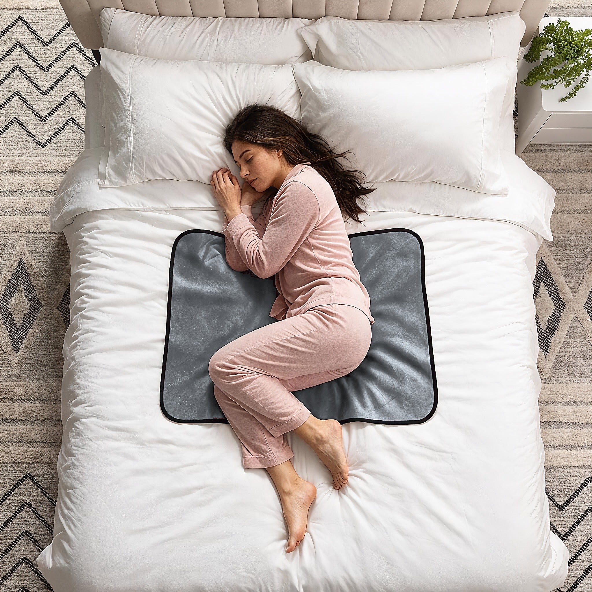 A woman in pink pajamas sleeps curled up on a white bed, resting on a dark gray rectangular mattress topper or pad with a black border. The scene is photographed from above, showing white pillows and bedding, a patterned rug, and a small plant in the corner.