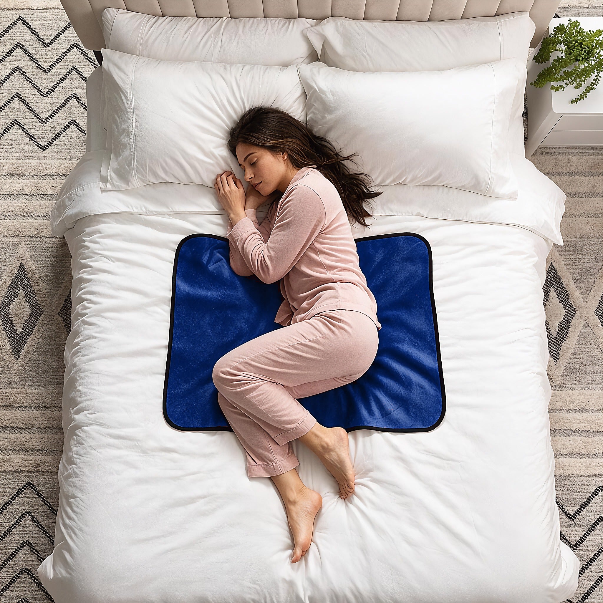 A woman in pink pajamas sleeps curled up on a white bed, resting on a blue contoured mattress topper.