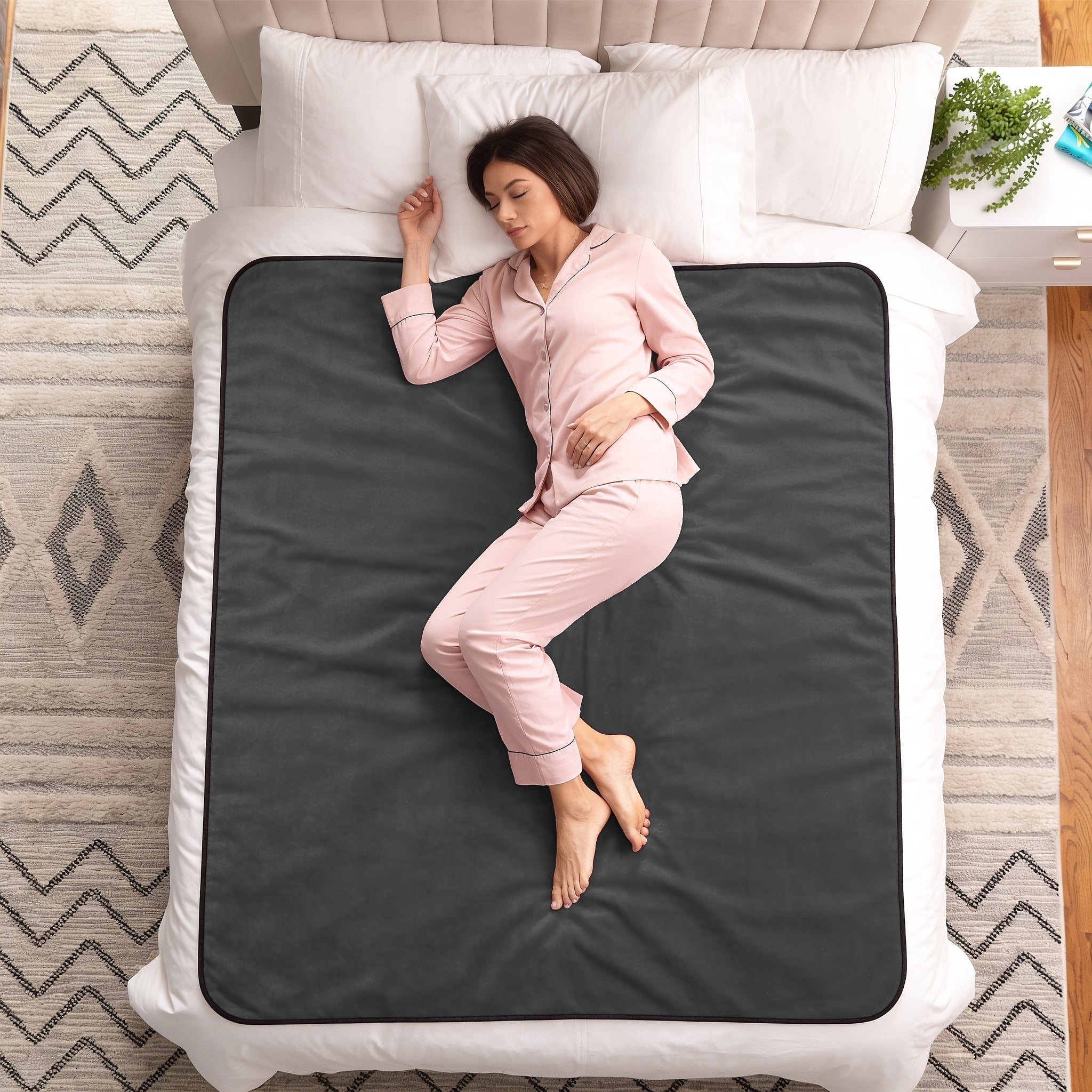 A woman in light pink pajamas lies on her side on a bed with a dark charcoal gray blanket or mattress pad underneath, surrounded by white bedding and pillows.