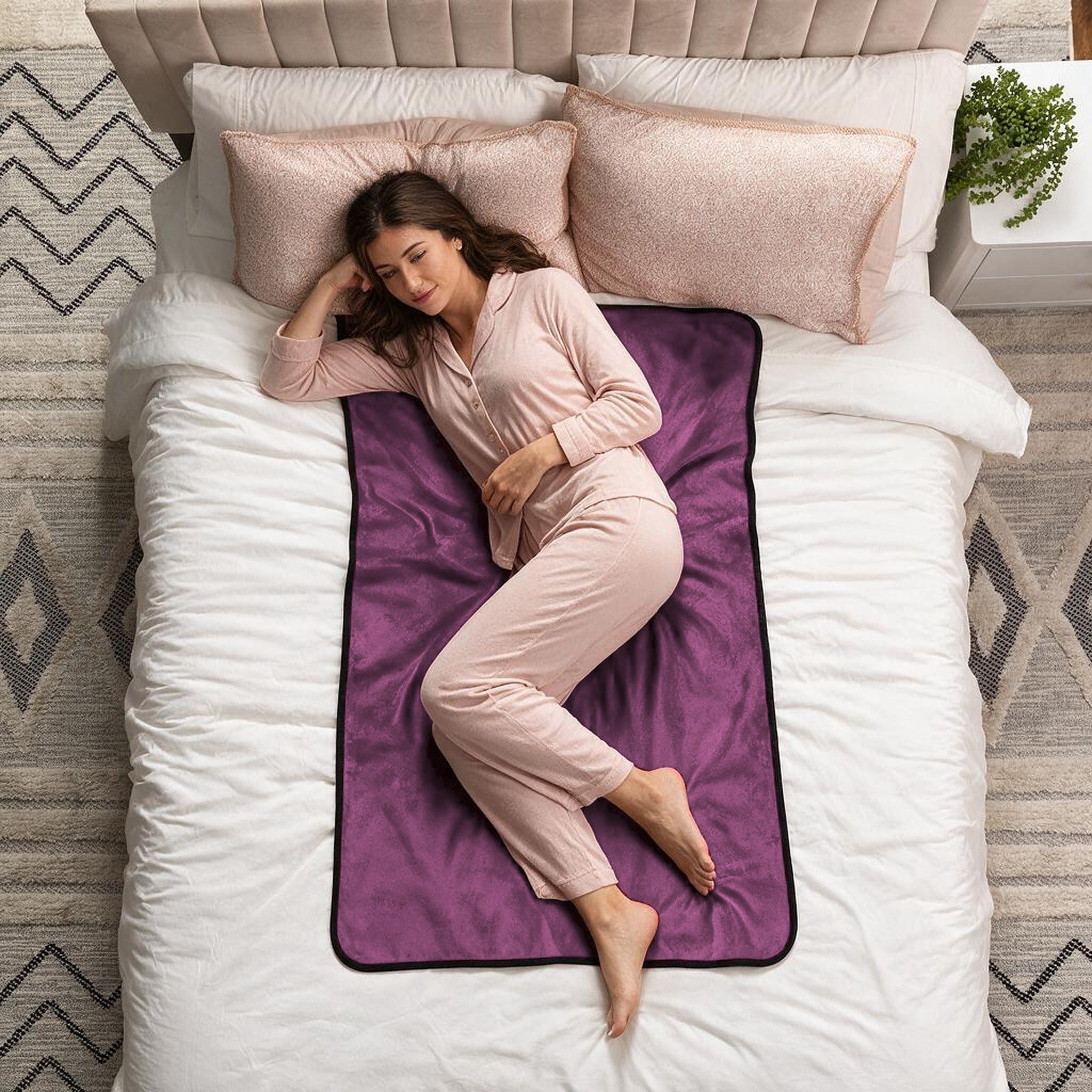 A woman in light pink pajamas lies on her side on a white bed, resting on a purple fleece blanket with a black border. The scene is styled as a top-down view with pillows, a headboard, and a patterned rug.