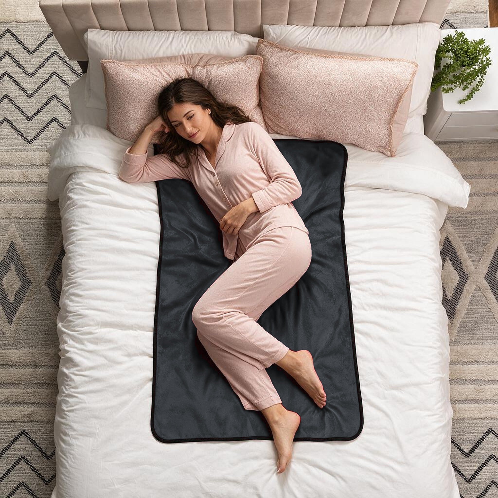 A woman in light pink pajamas lies on her side on a bed with white bedding and a dark charcoal fleece blanket underneath her torso. The bed has blush pink pillows, and the room is styled with a patterned rug and a small plant on a side table.