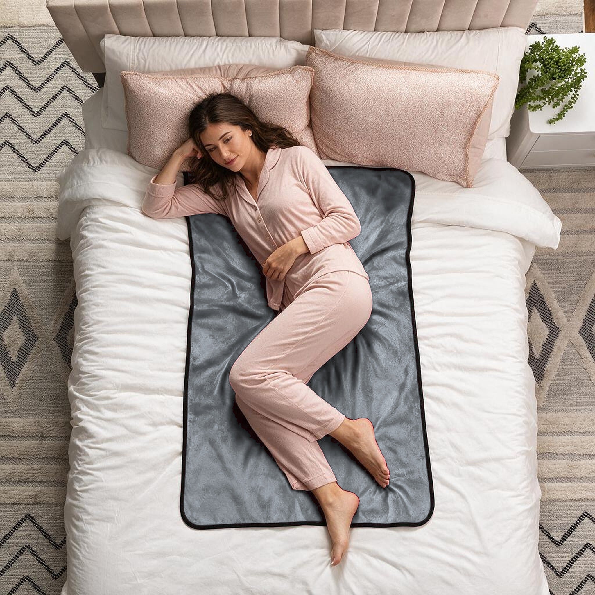 A woman in light pink pajamas lies on her side on a bed with white bedding and pink pillows, using a dark gray blanket with a black border as a support cushion. The scene is shot from a top-down view in a styled bedroom setting.
