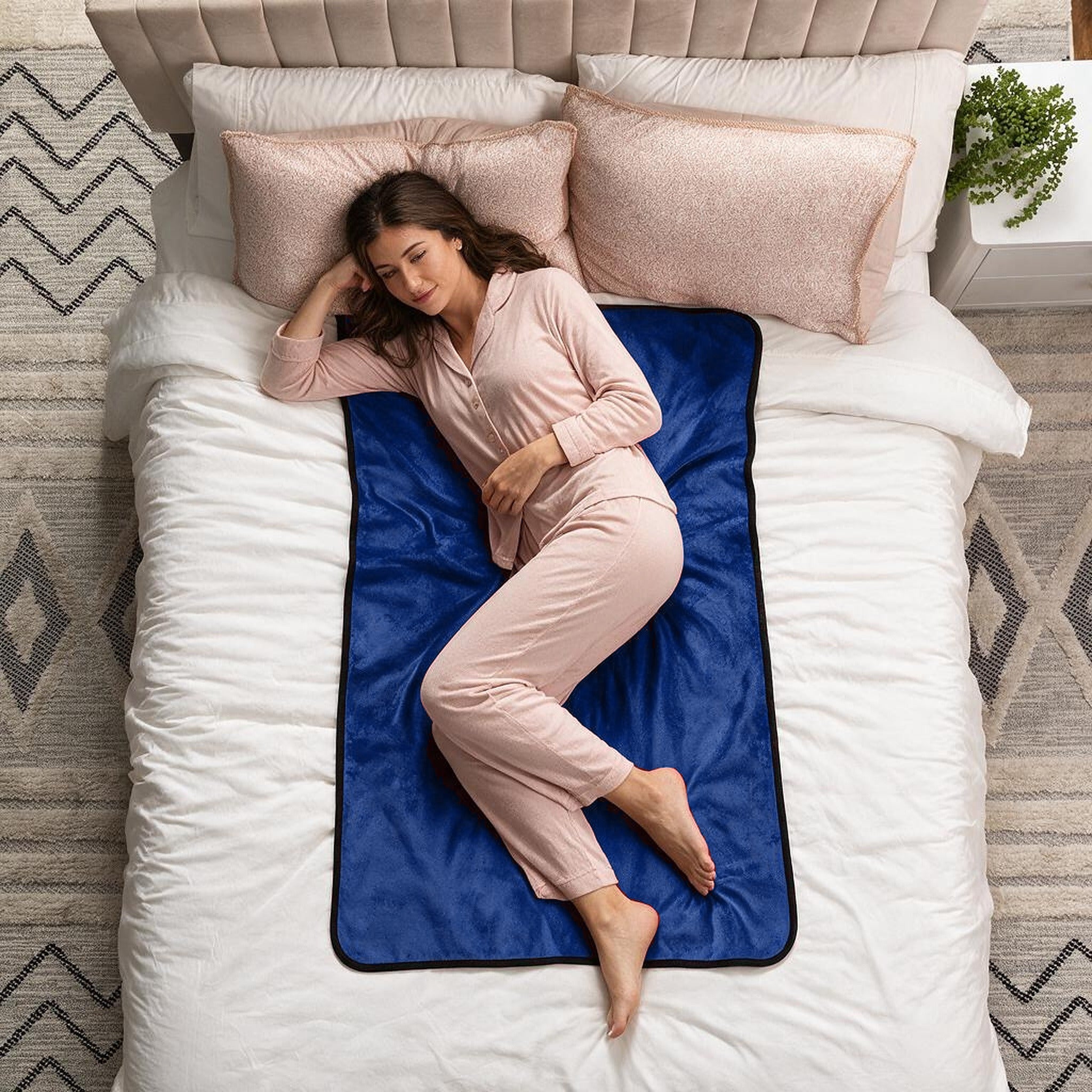 A woman in light pink pajamas lies on her side on a bed with white bedding, pink pillows, and a dark blue blanket or mat spread beneath her.