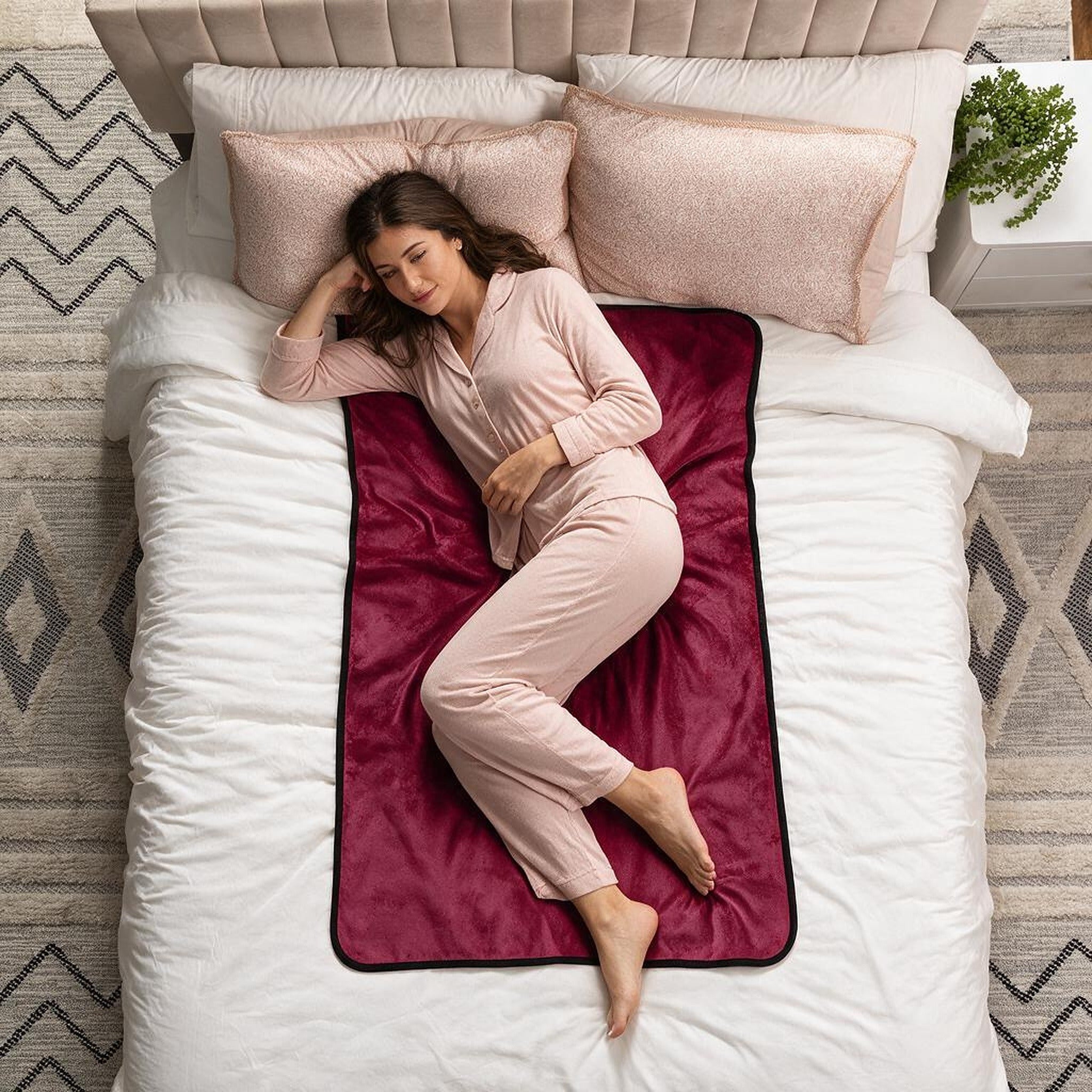 A woman in light pink pajamas lies on her side on a bed with white bedding, a burgundy fleece blanket with a black border, and blush pink pillows.