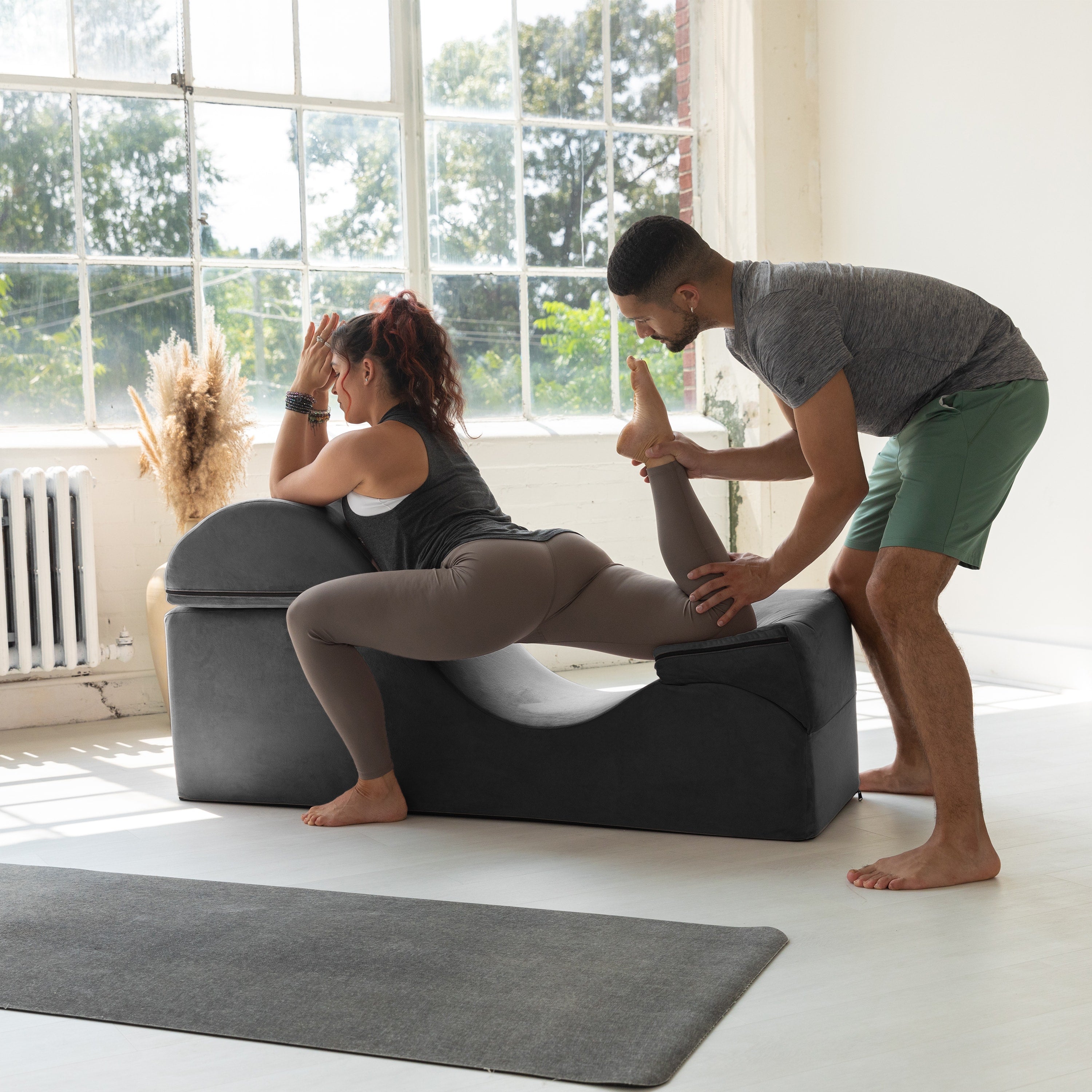 Person assisting another person with a yoga pose on a gray cushion in a bright room.