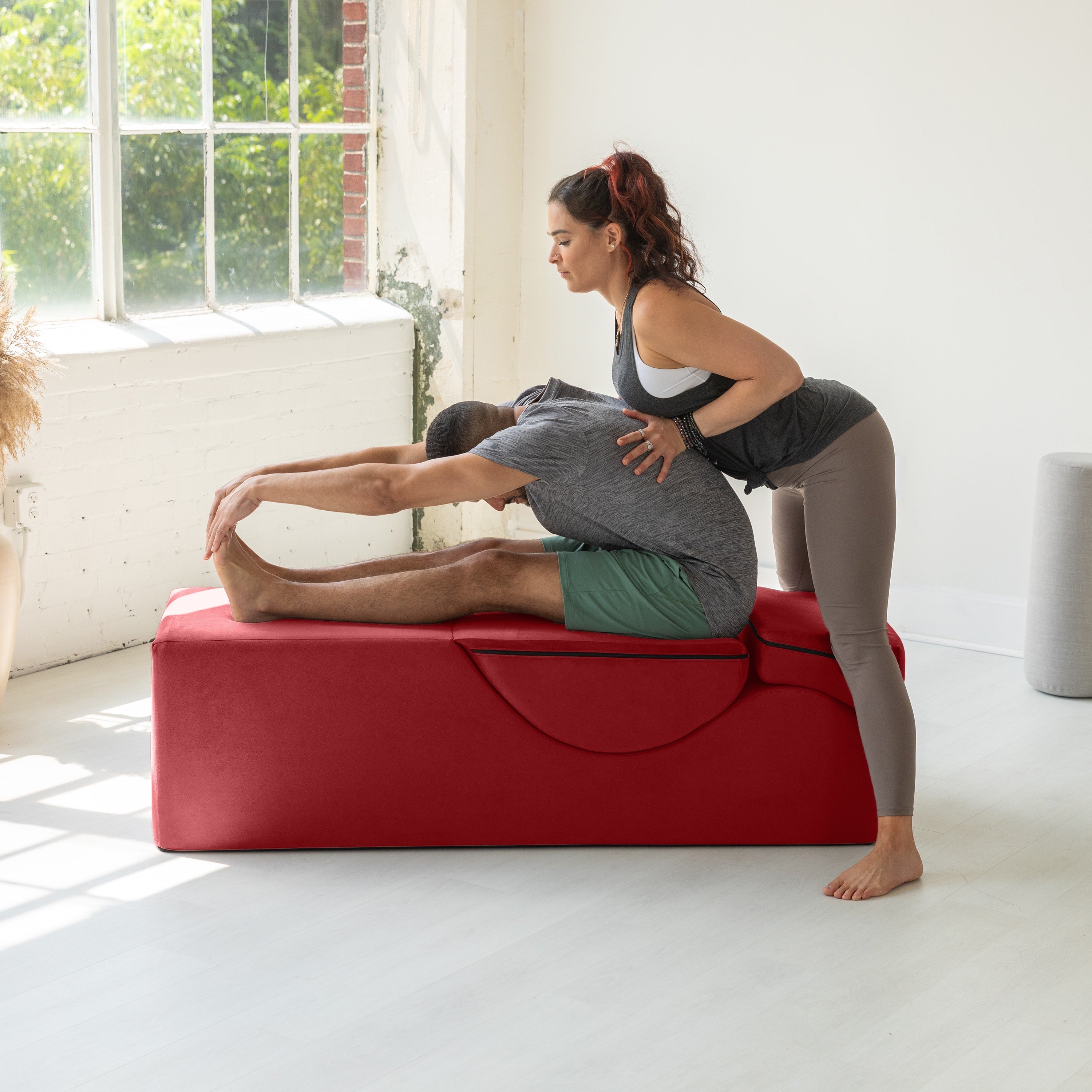 Two women exercising on a red fitness bench in a bright room with large windows.