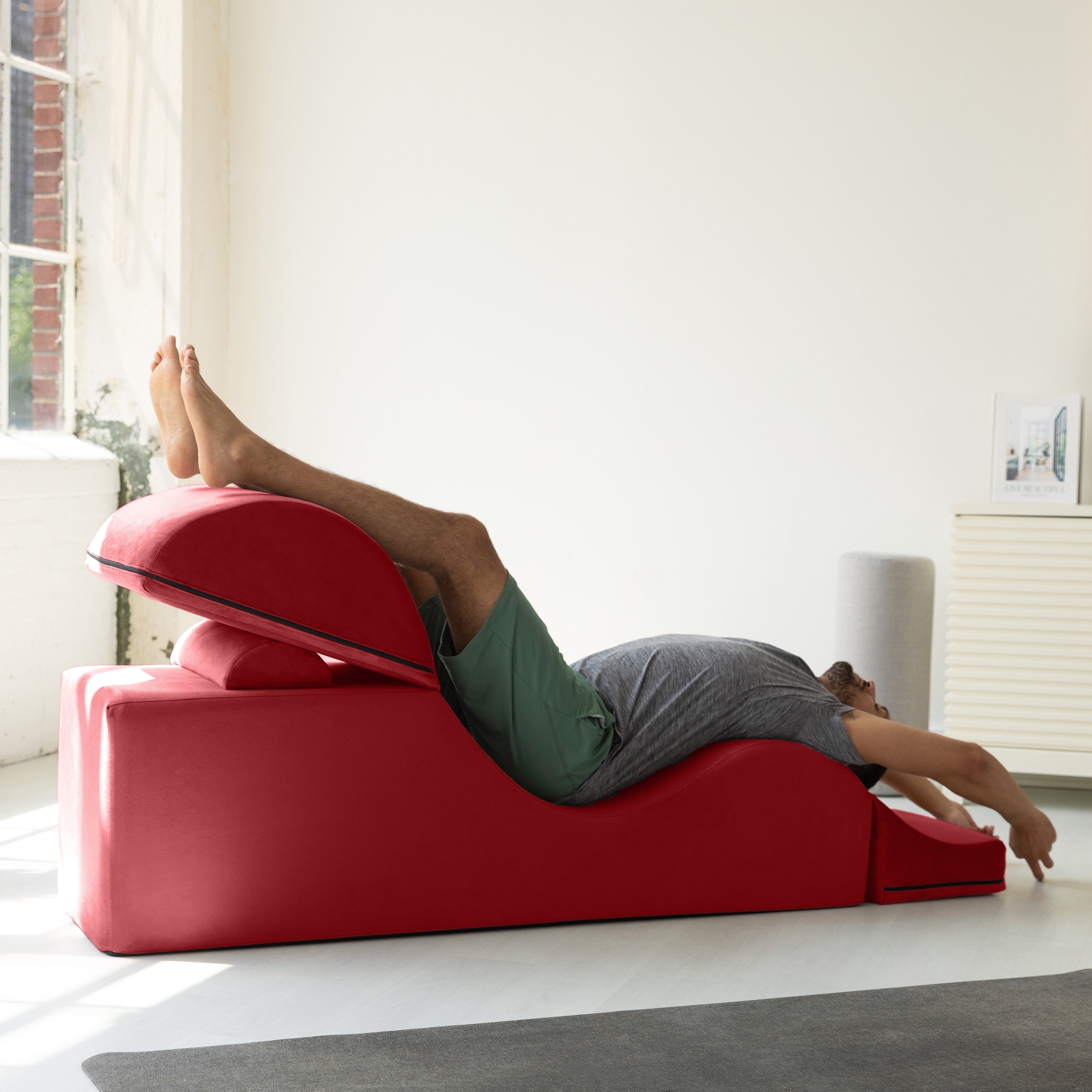 A person lying on a red contoured massage or therapy table in a bright, modern room with white walls and a window.