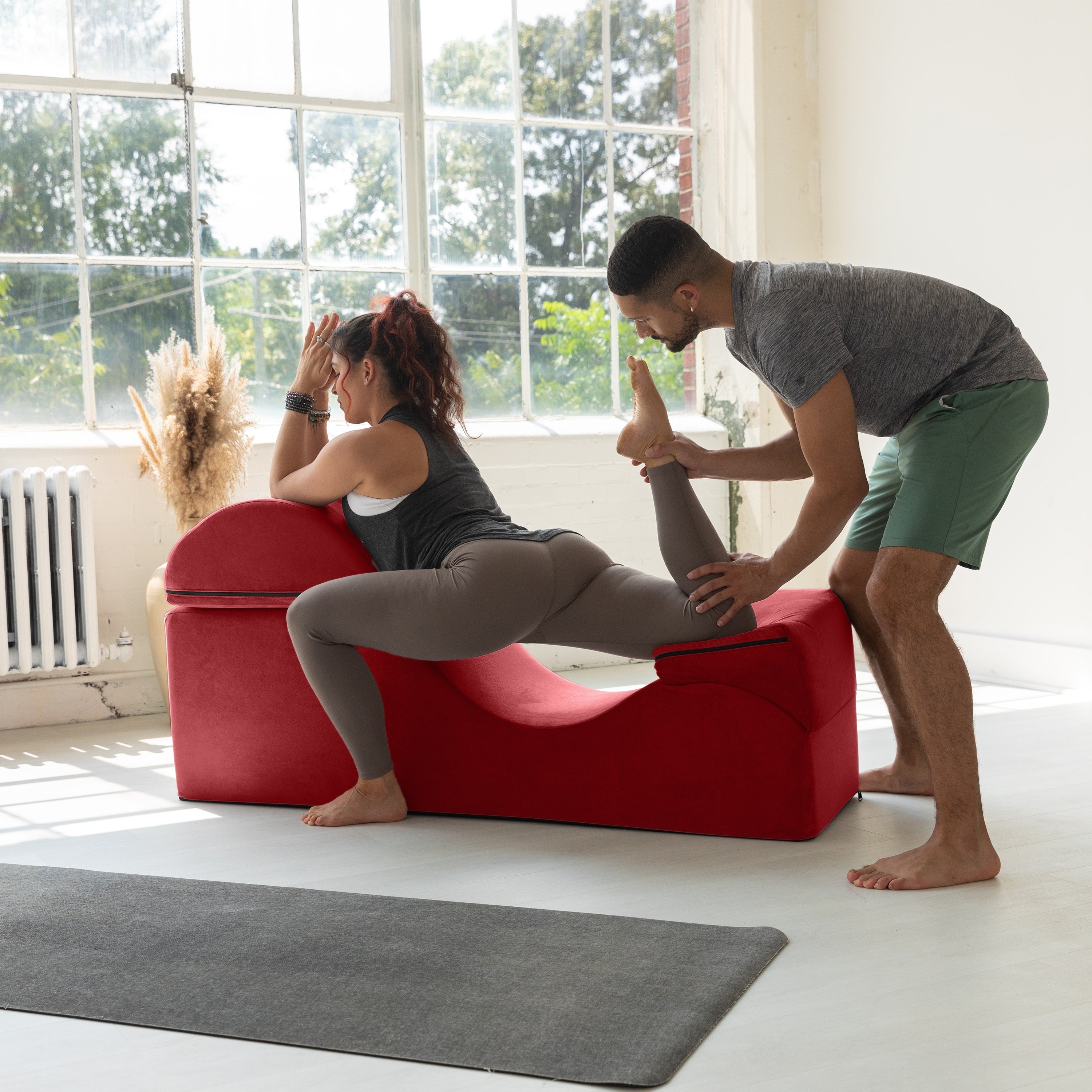Person using a red exercise machine with another person assisting them in a bright room.